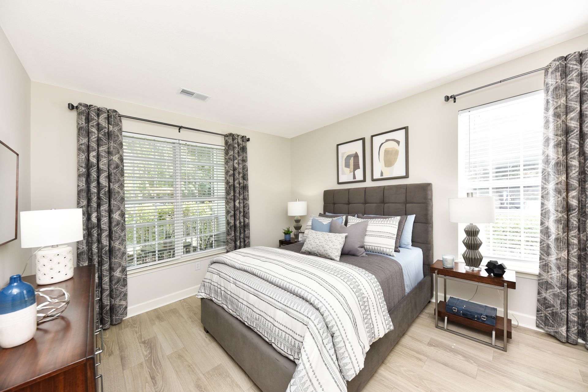Bedroom with bed, curtains, nightstands, dresser, and windows. Light gray walls and wood flooring.