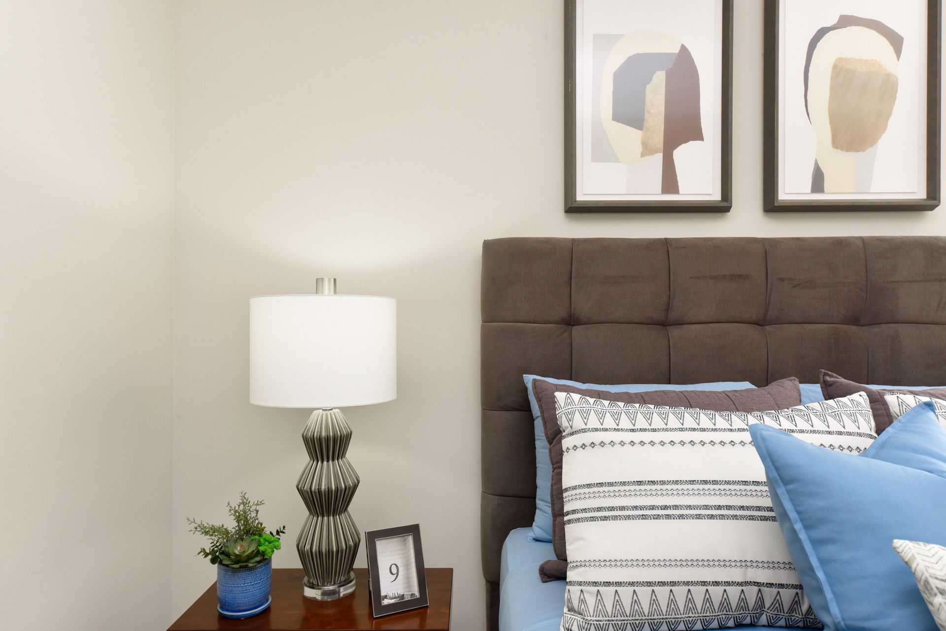 Bedroom corner with a bed, lamp, artwork, and nightstand. Light blue and brown color scheme.