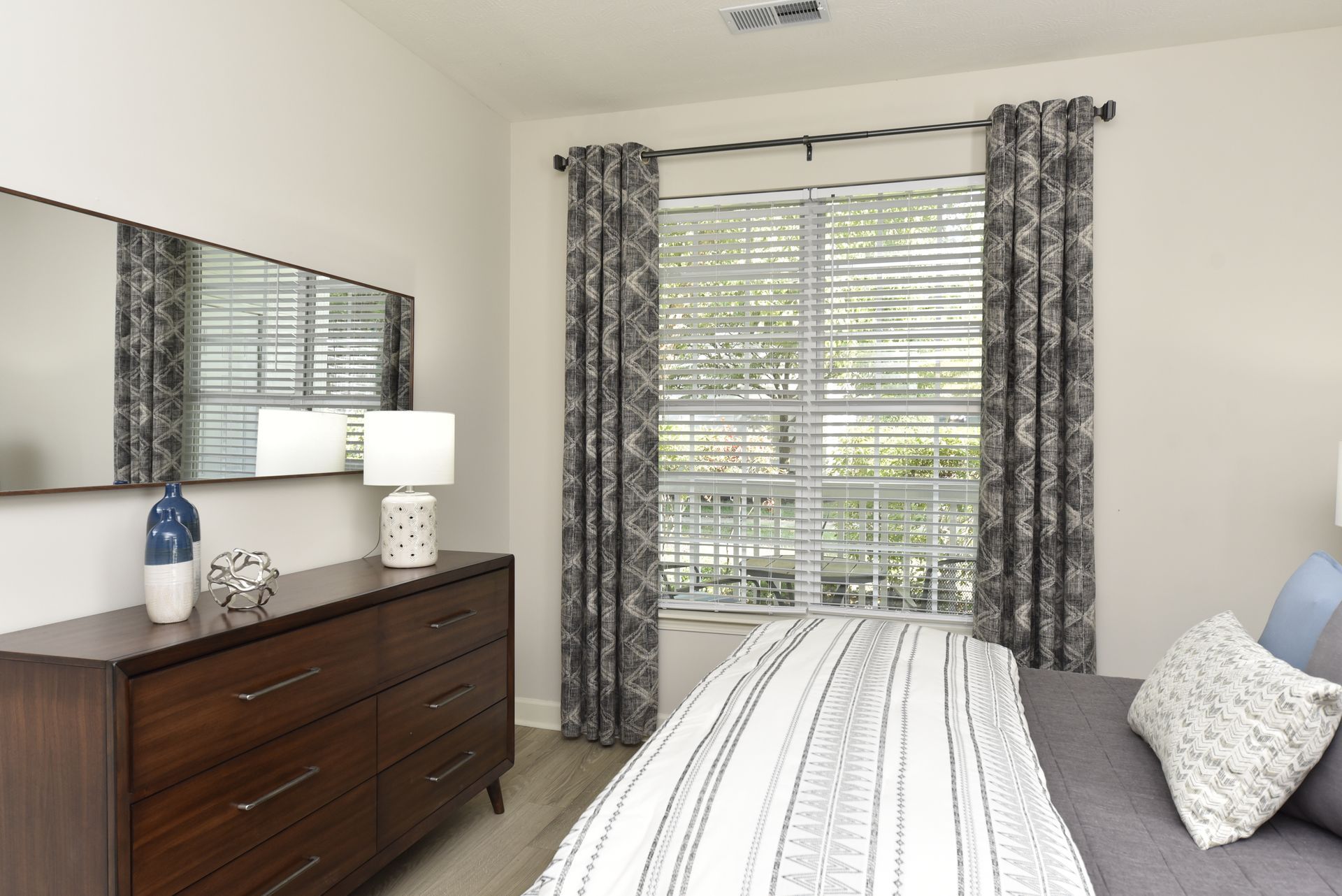 Bedroom with dresser, mirror, window, patterned curtains, and bed with pillows.