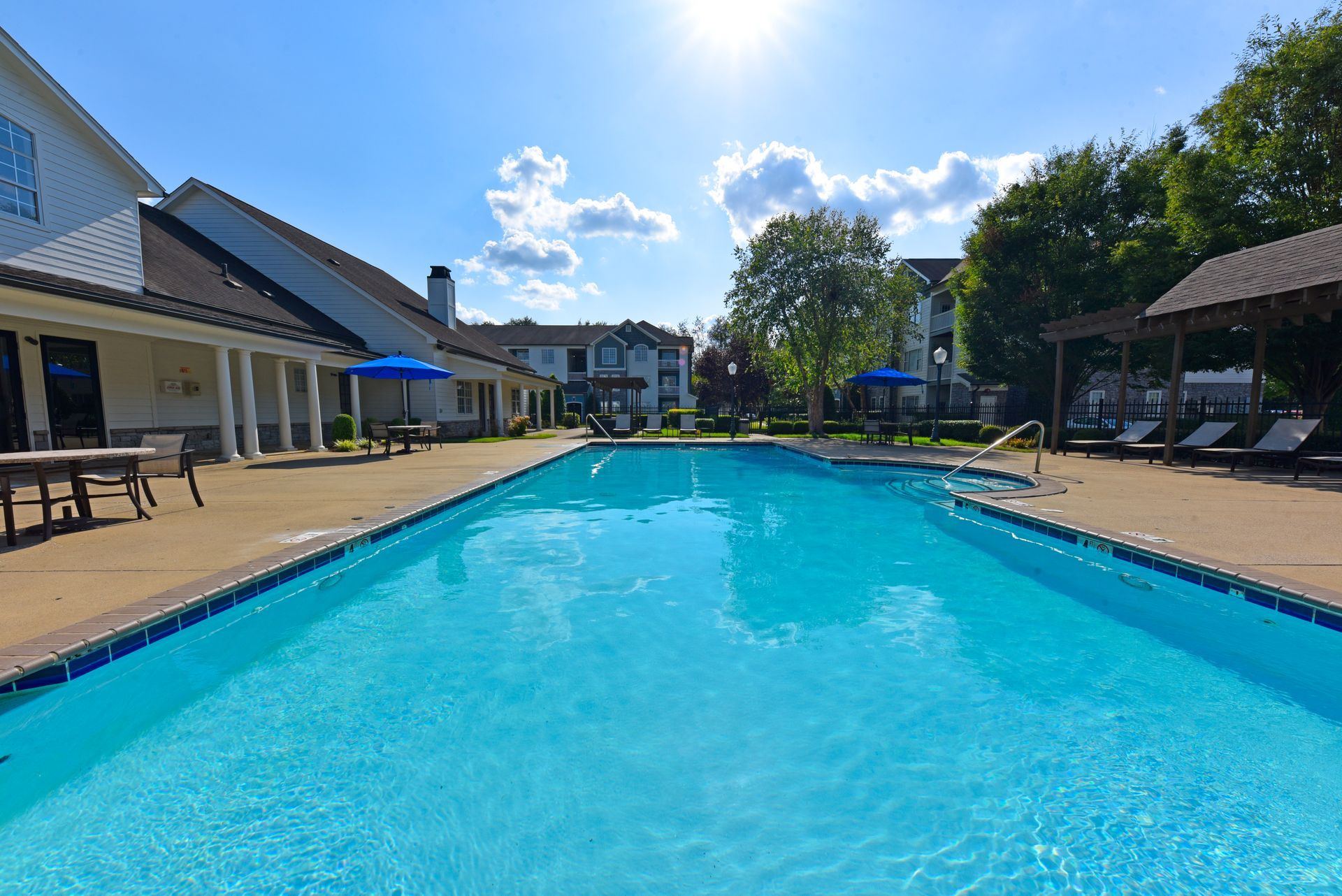 Swimming pool in a courtyard, sunny day. Buildings surround, blue umbrellas, and people relaxing by the water.