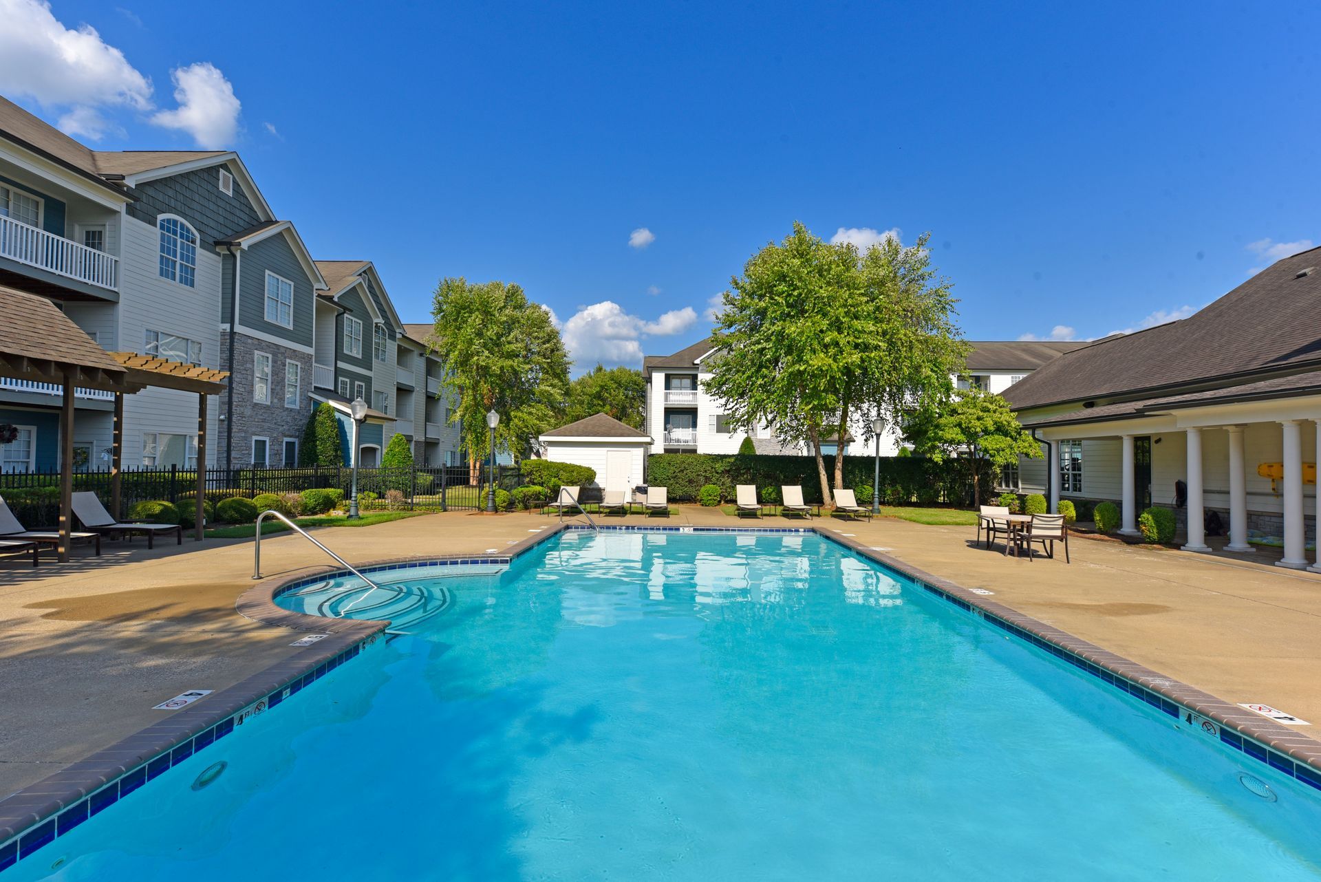 Swimming pool in apartment complex courtyard on a sunny day. Blue water, lounge chairs, and buildings are visible.