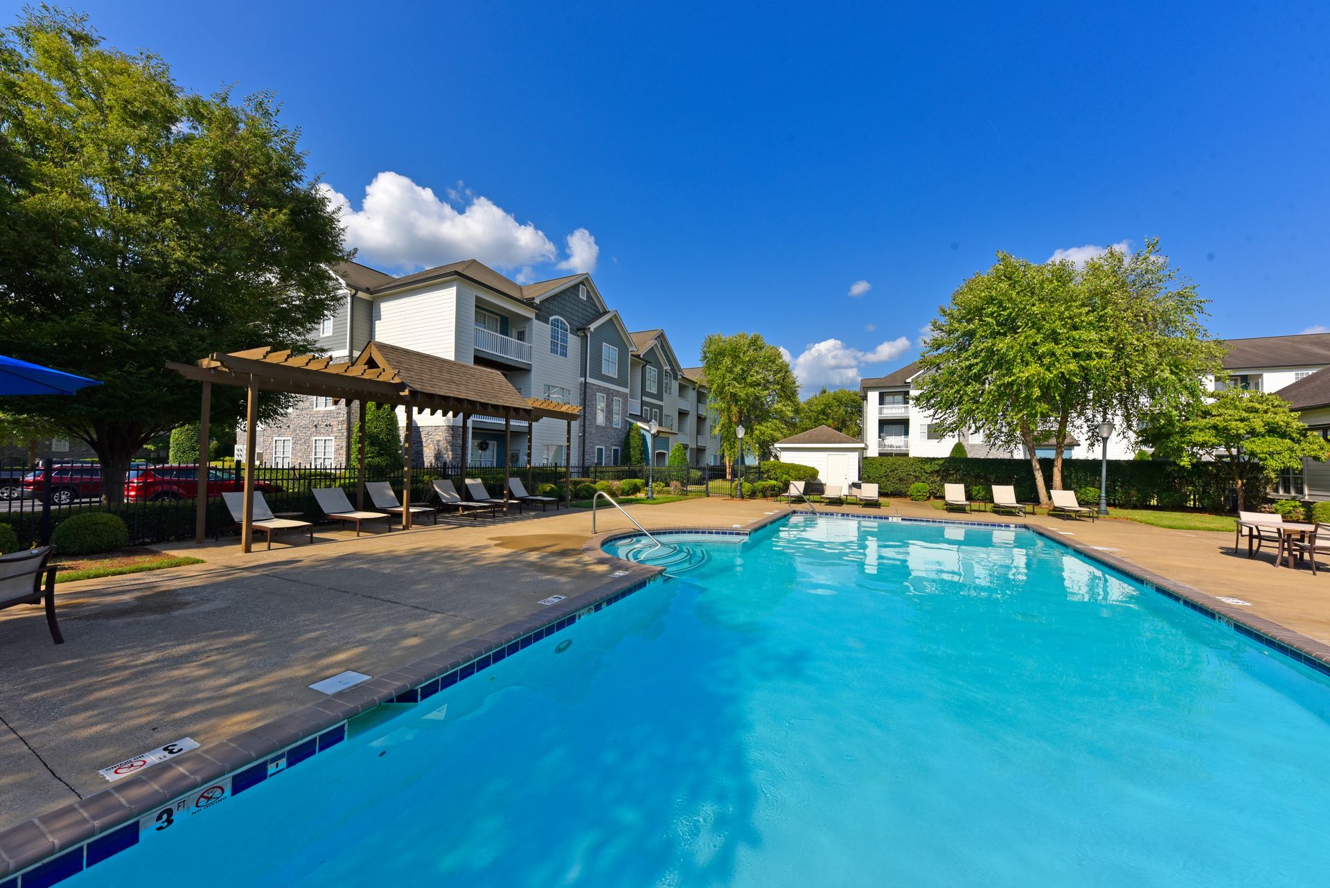 Pool with blue water, lounge chairs, and a multi-story apartment building under a blue sky.