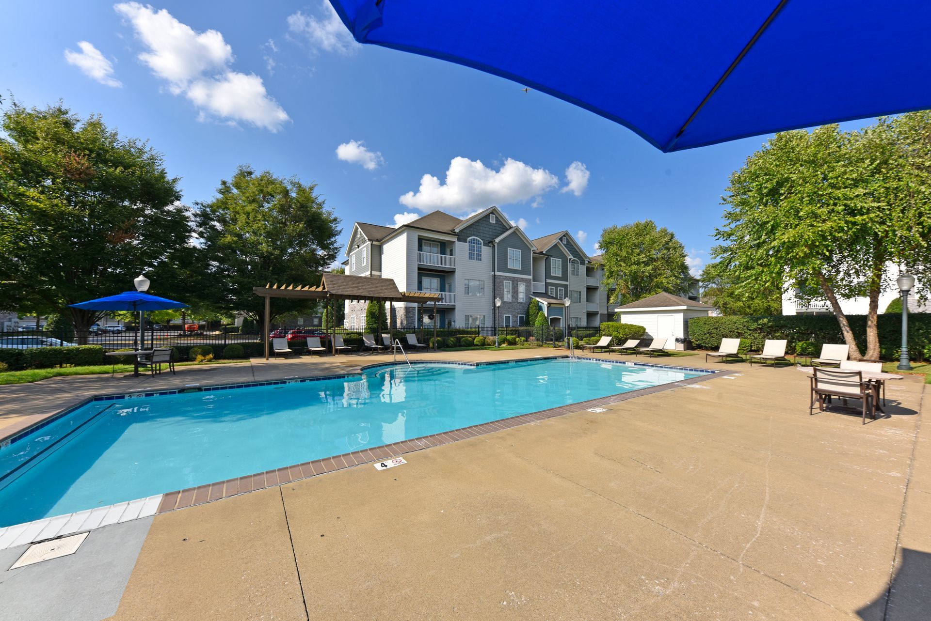 Pool with lounge chairs and apartment building under a blue sky.