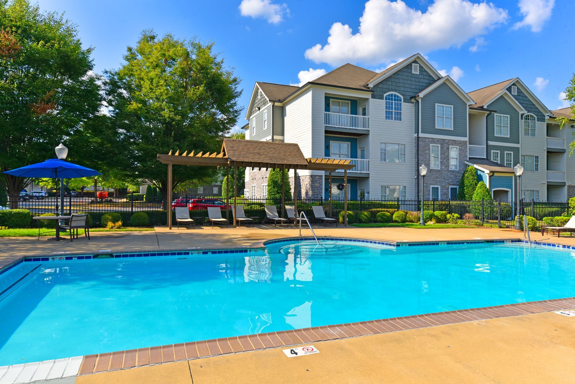 Apartment complex with pool, lounge chairs, and pergola under a blue sky.