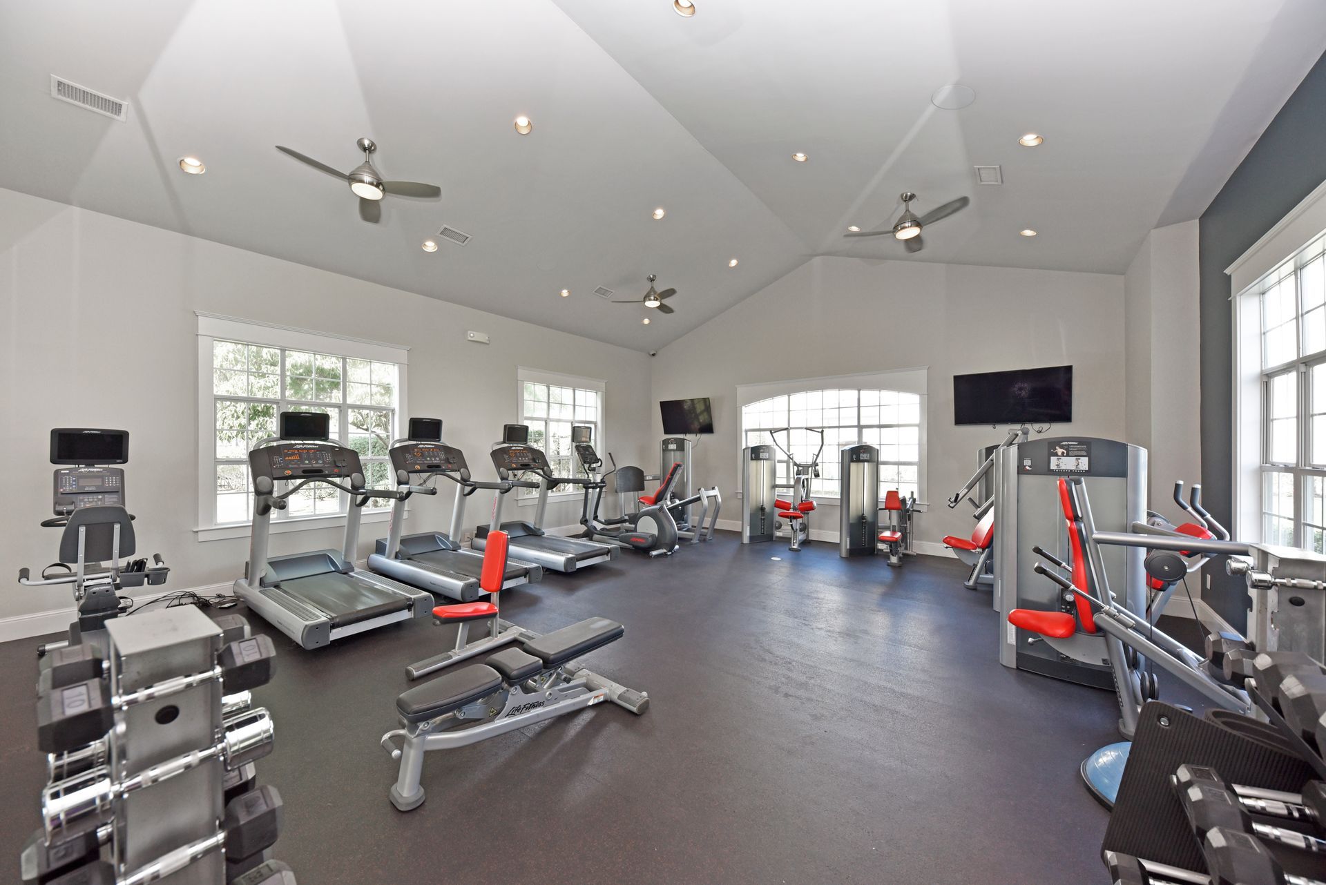 Gym with exercise machines, weights, windows, ceiling fans, and TVs.