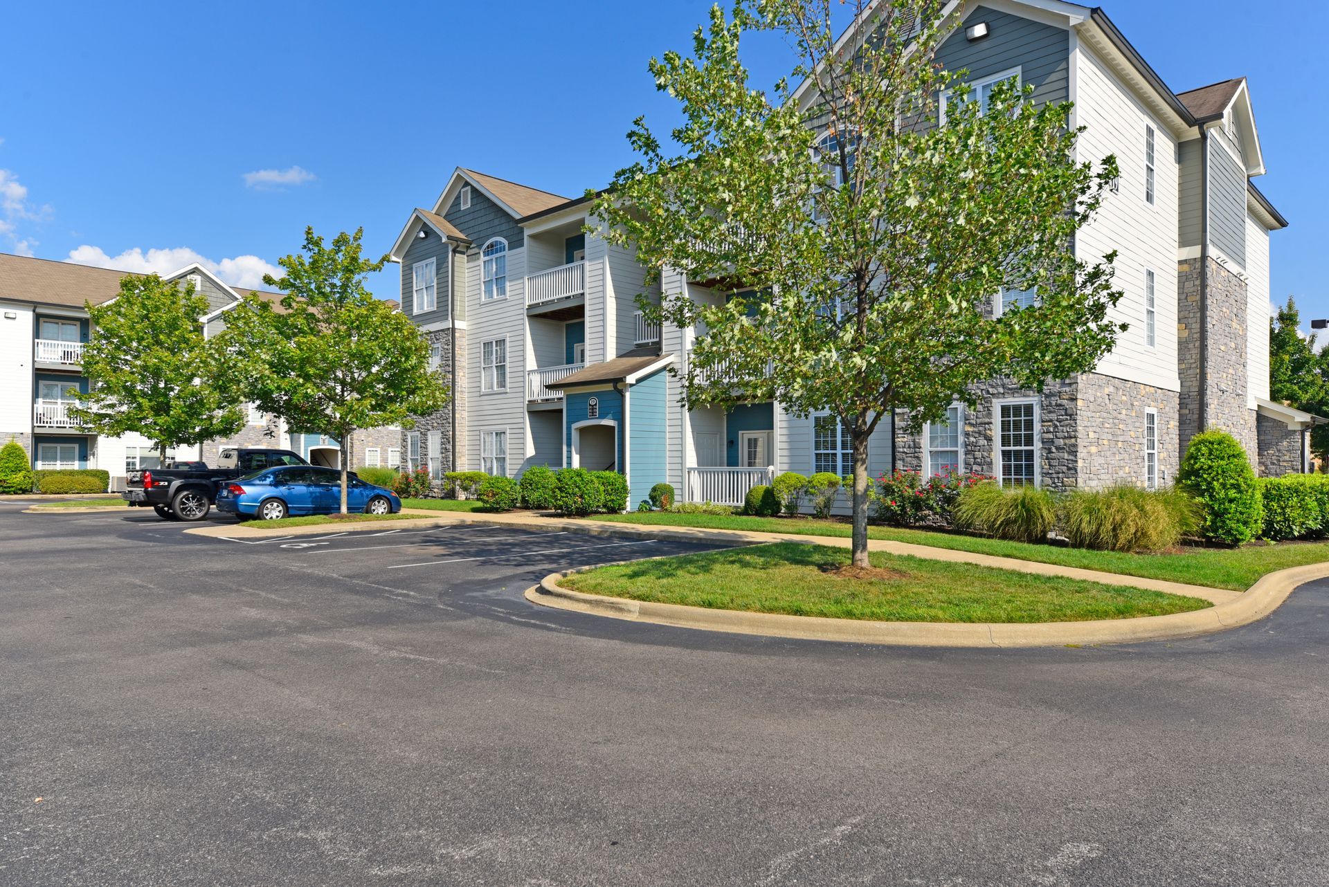 Apartment complex with parking lot, blue sky, and green trees.