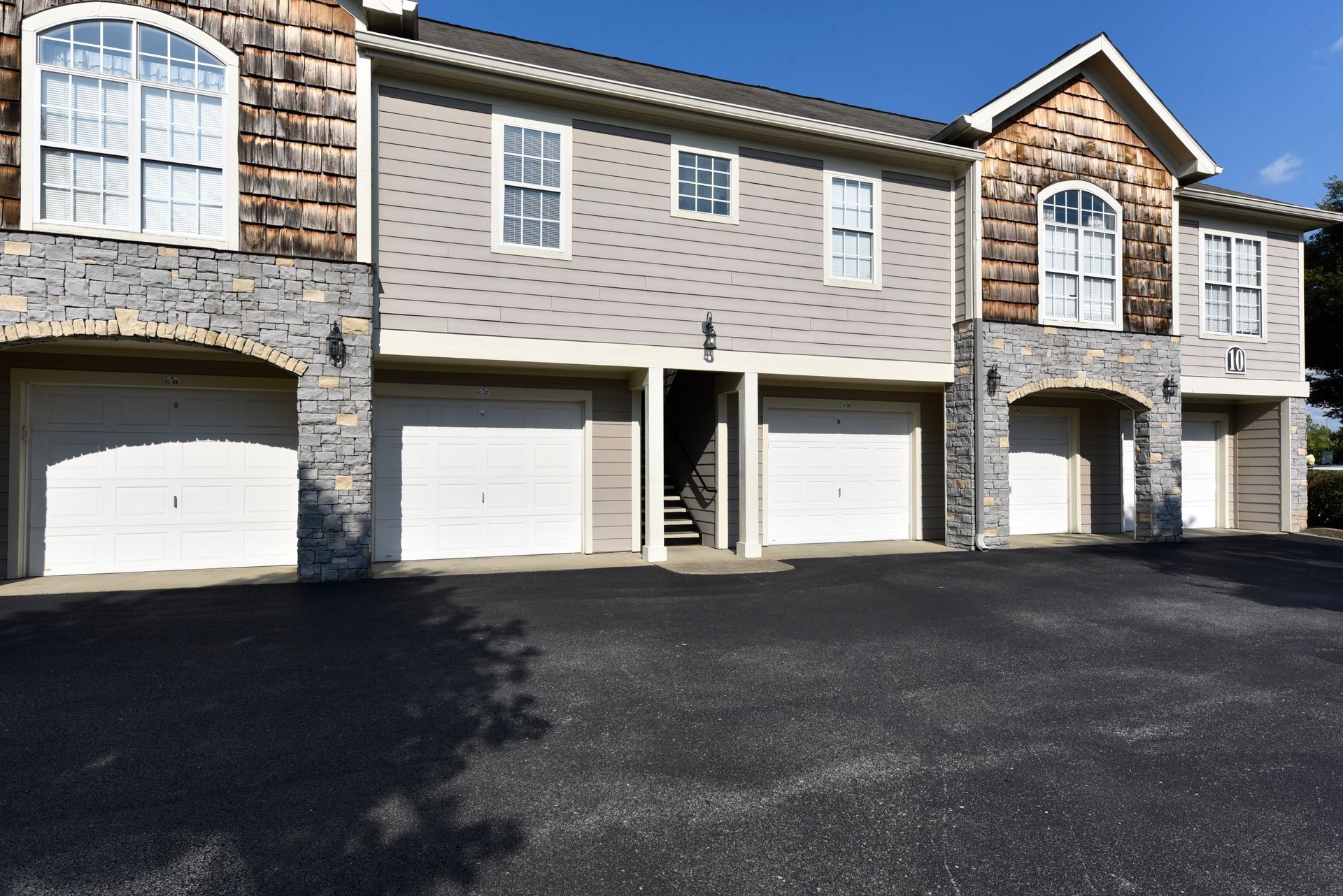 Apartment building with garages. Stone and gray siding, white garage doors, paved driveway. Sunny day.
