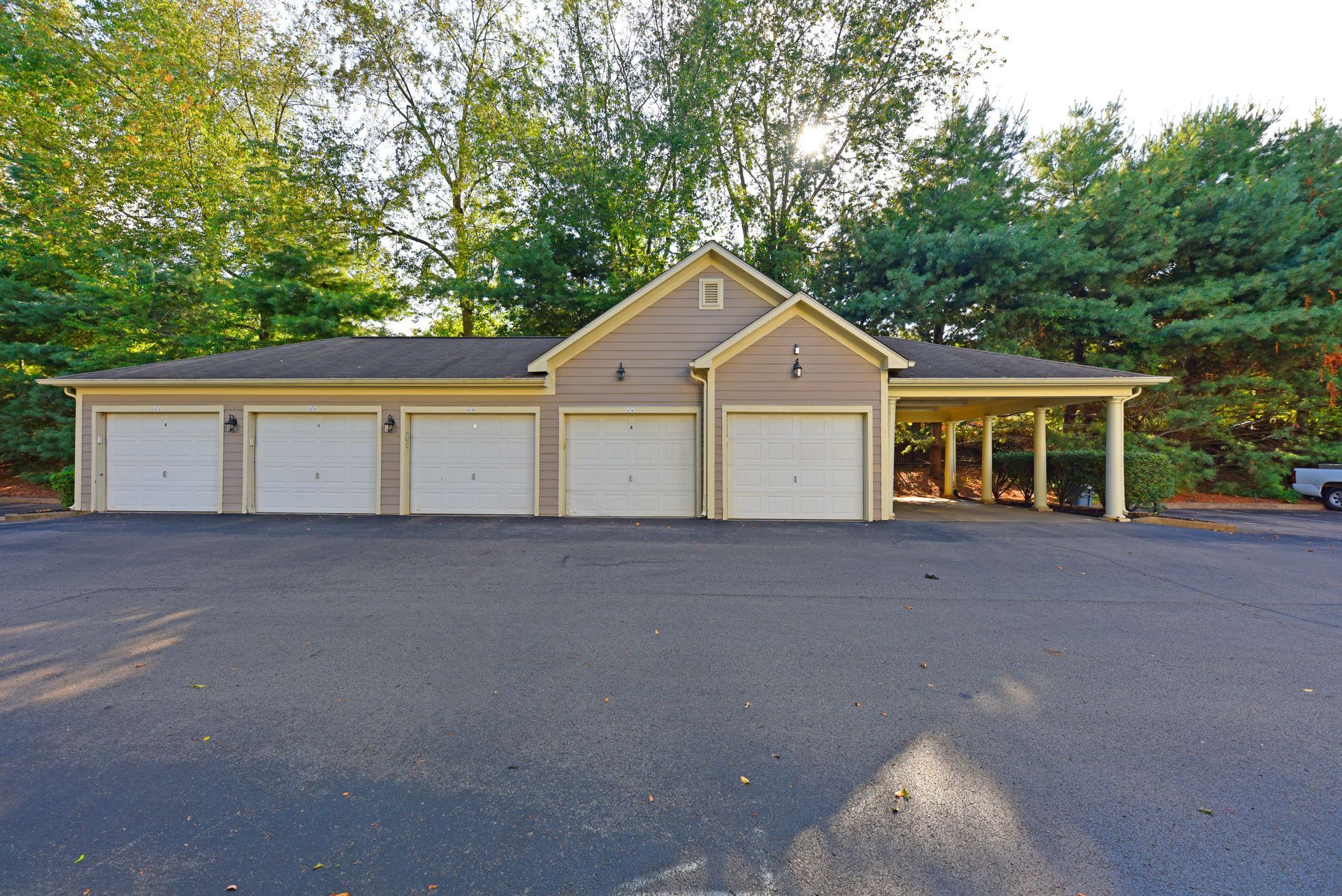 Five-car garage with beige doors, brown roof, and carport, set against a backdrop of trees and asphalt.