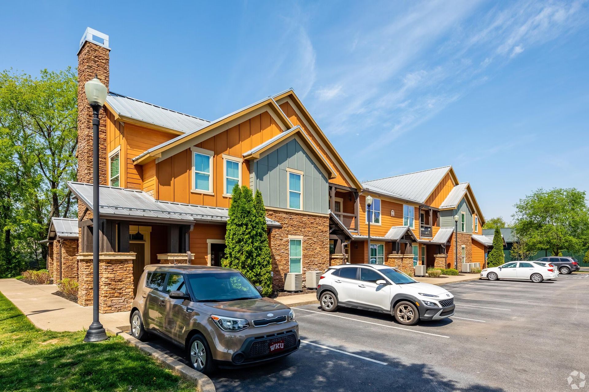 Apartment complex with brown siding, stone accents, and parked cars under a blue sky.