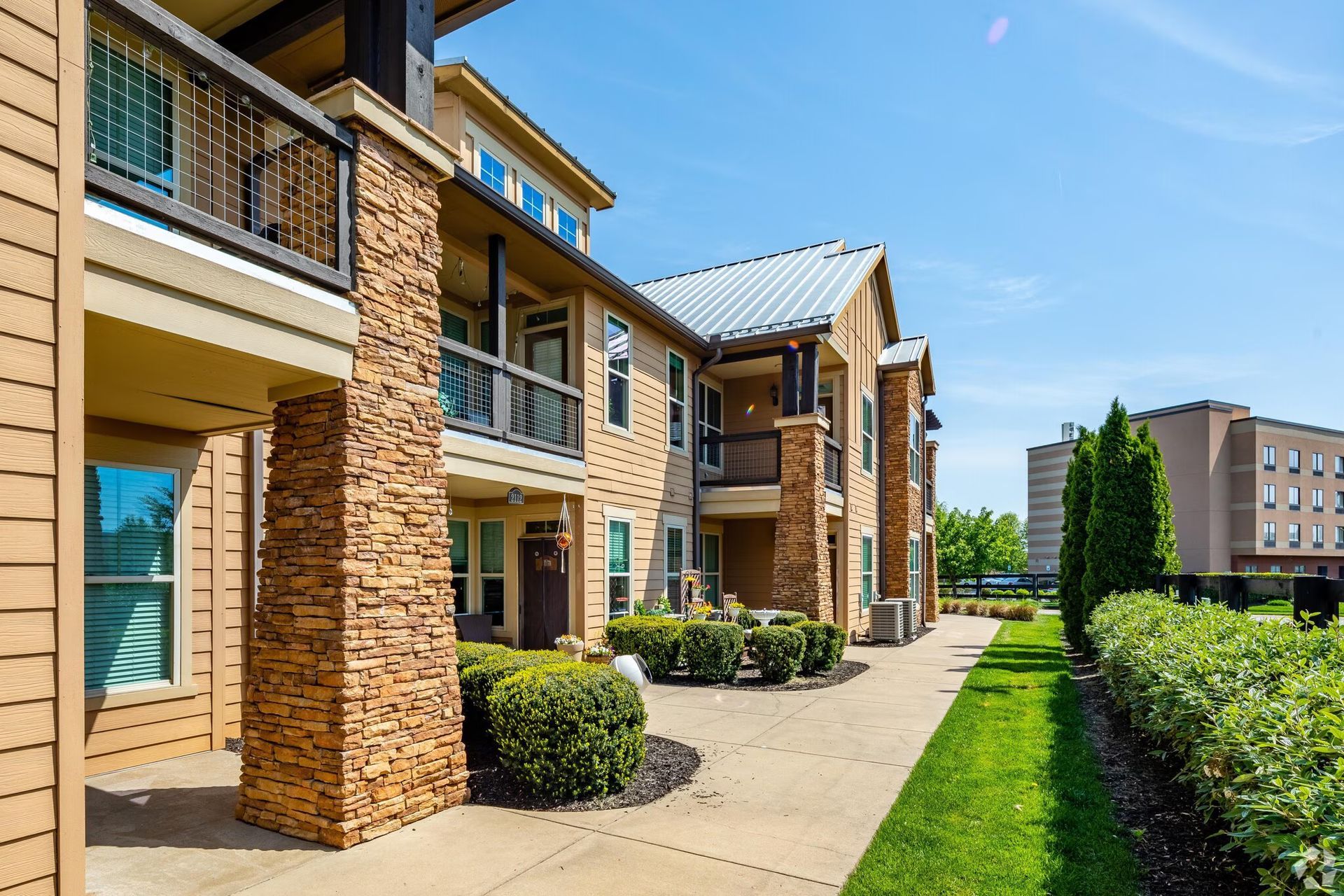 Apartment complex exterior with stone columns, balconies, and walkway. Green grass and shrubs.
