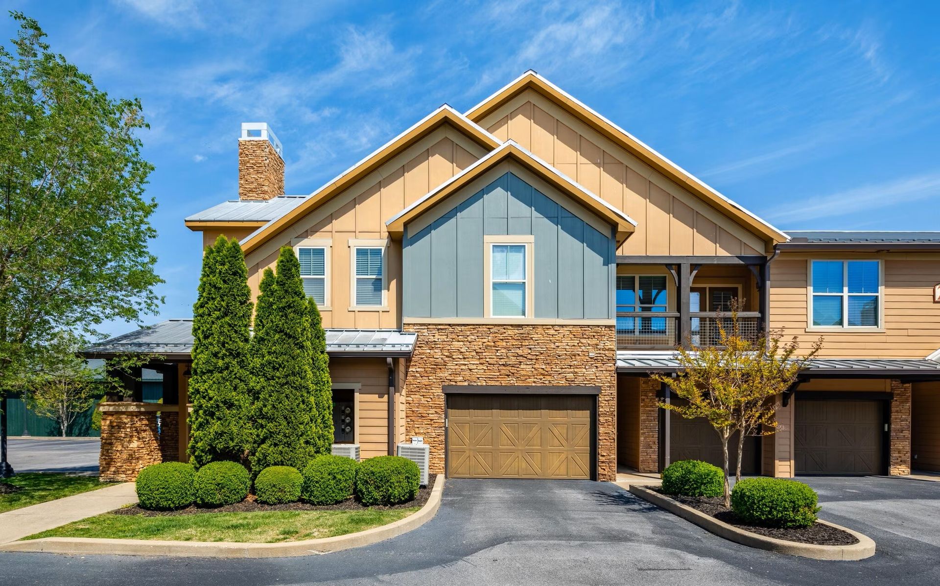 Townhouses with tan siding, brown garage doors, blue accents, and manicured landscaping on a sunny day.