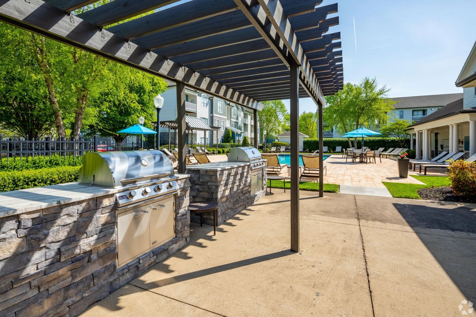Outdoor grilling area with stone counters, shaded by a pergola, overlooking a pool.