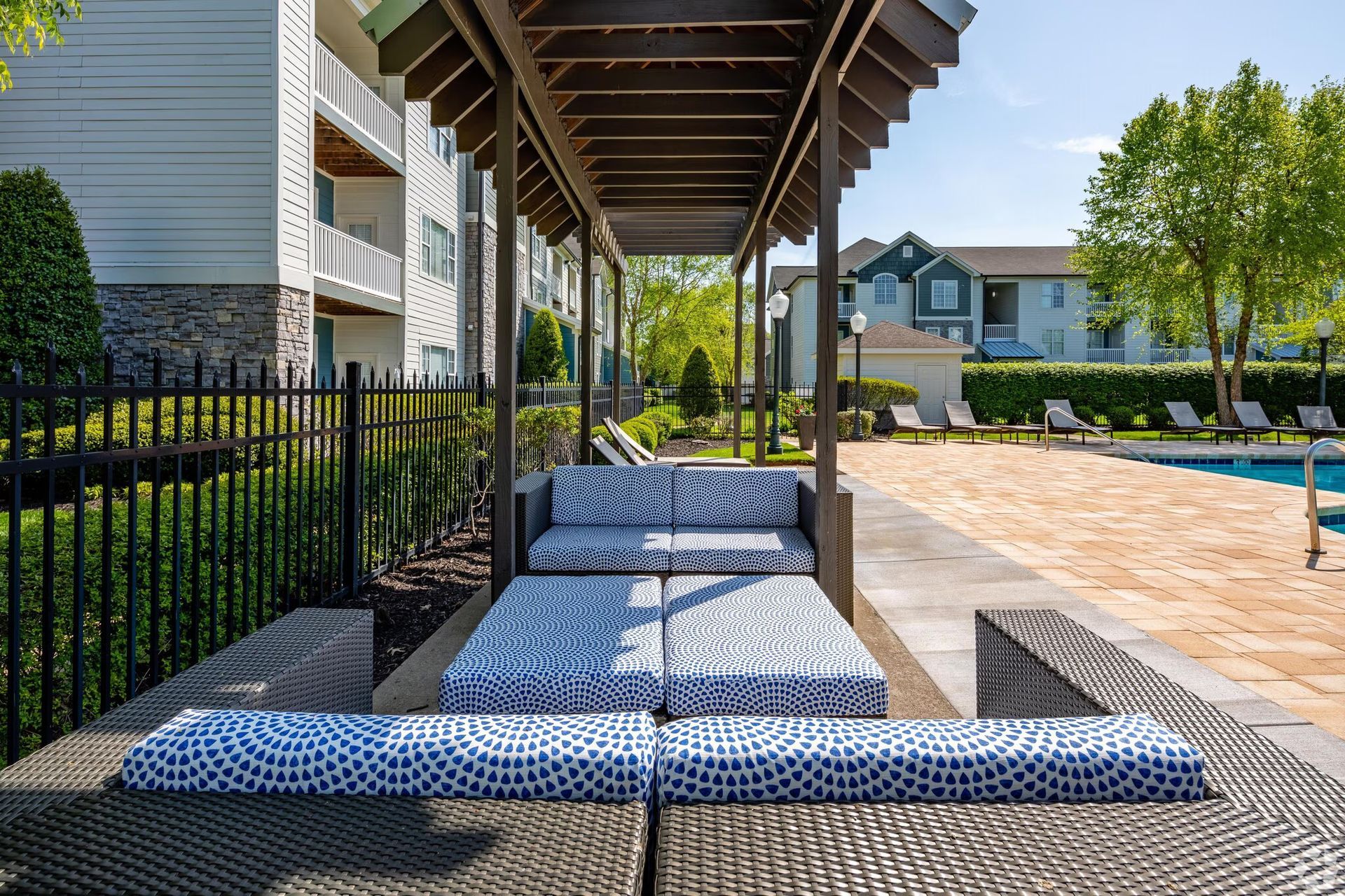 Outdoor lounge area with cushioned seating under a wooden canopy next to a pool.