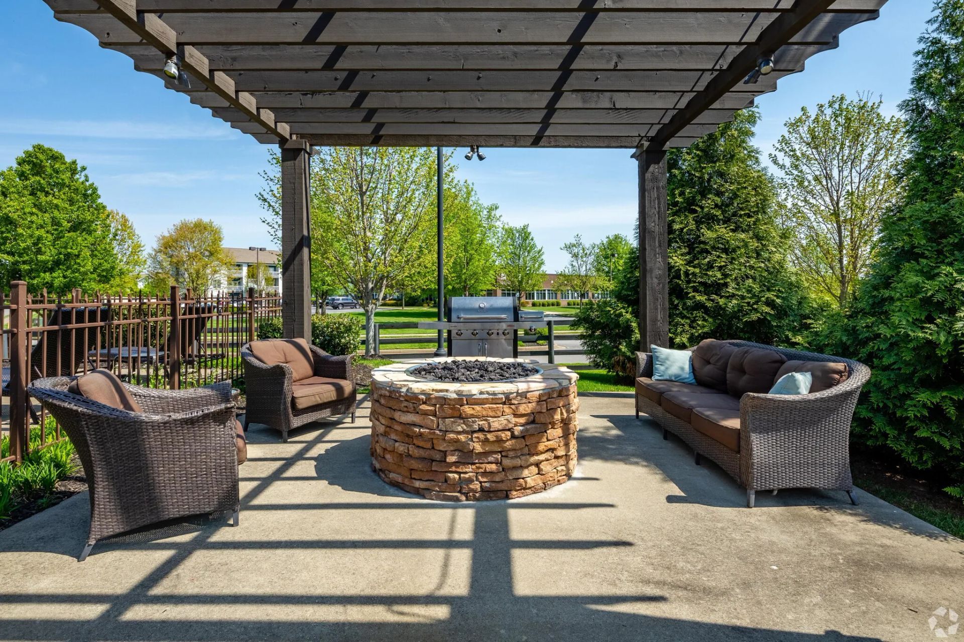 Outdoor patio with seating around a stone fire pit, under a pergola. Grill in the background.