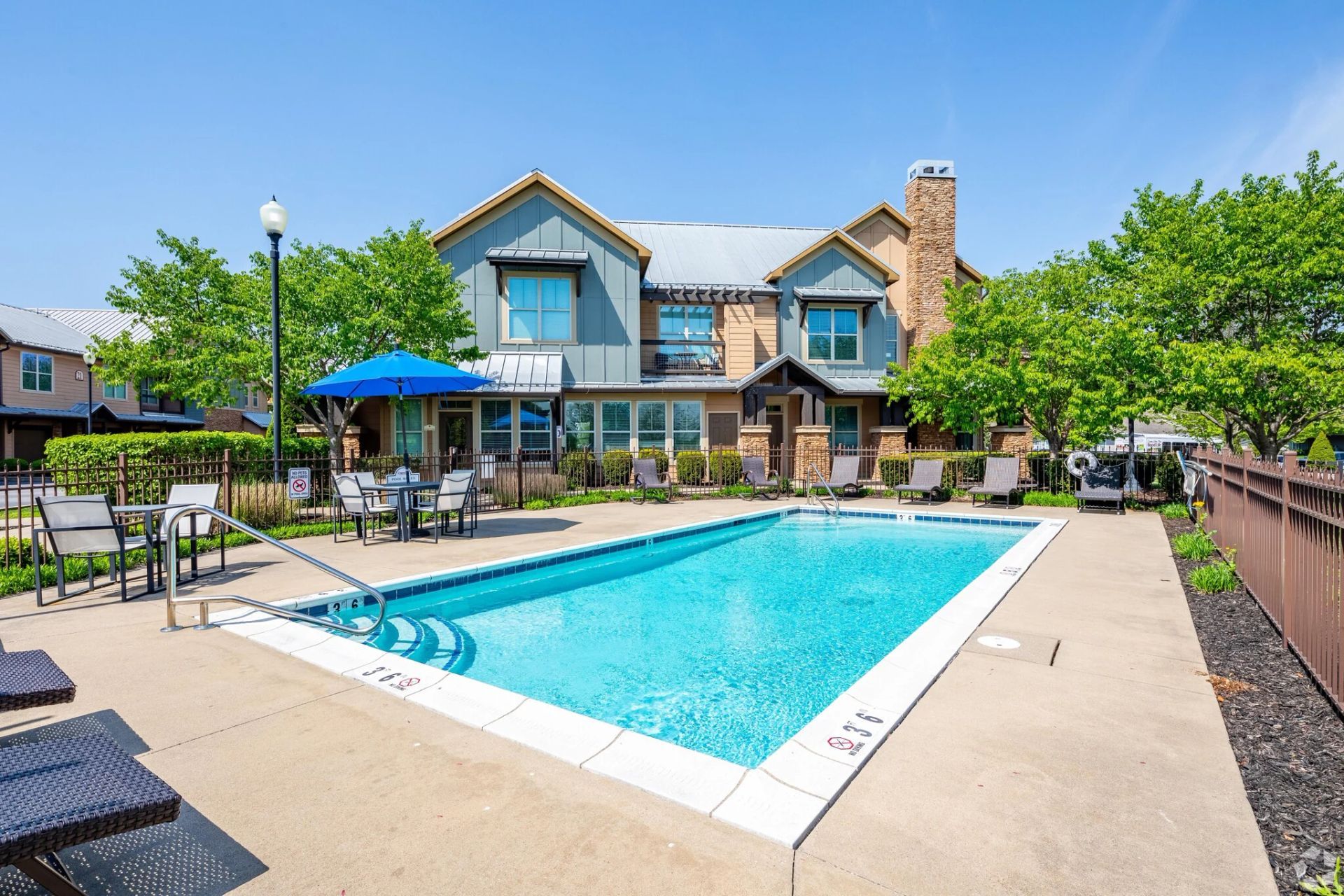 Swimming pool in front of a two-story building with outdoor seating and blue umbrella; sunny day.