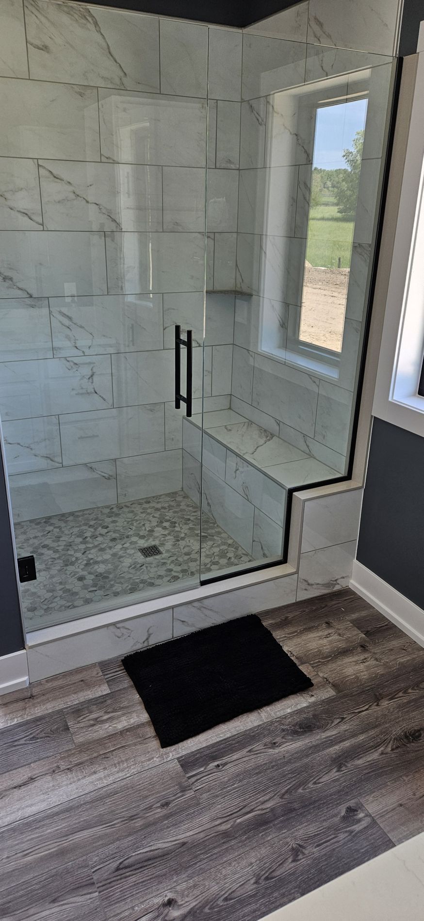 Modern shower with white tiled walls and glass door, bench seat, and black handle.