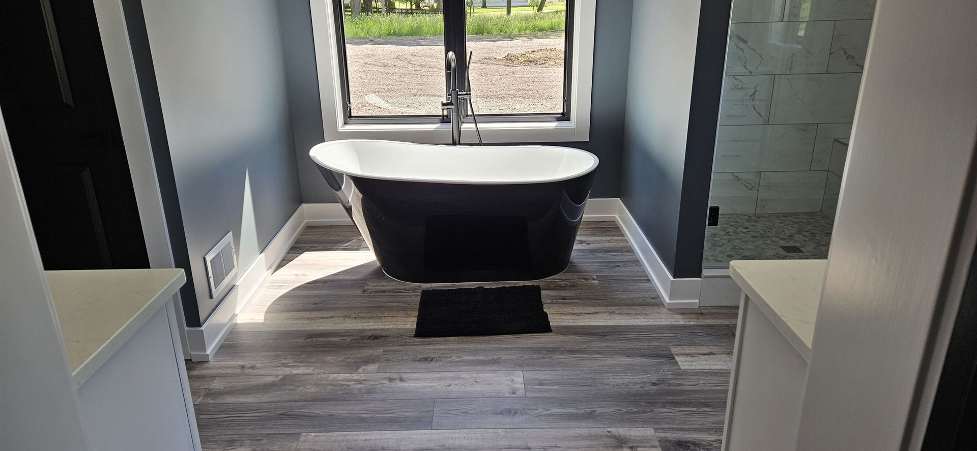 Bathroom with black freestanding tub, gray walls, and wood-look flooring. Window behind the tub.