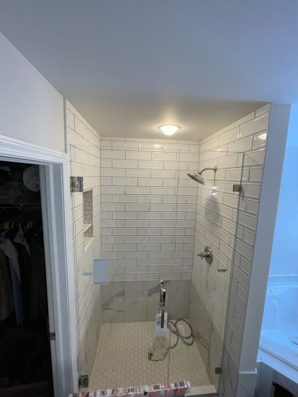 A small, white-tiled shower with glass doors, a shower head, and a handheld wand, next to a closet.