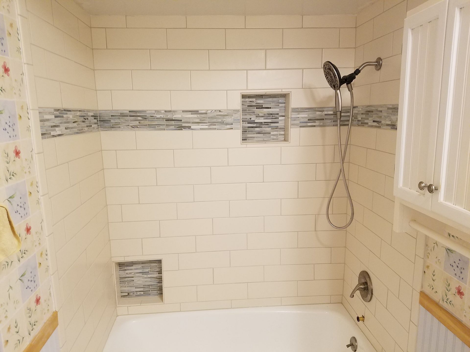 Bathroom shower with cream-colored tiled walls, a decorative tile strip, a showerhead, and two recessed shelves.