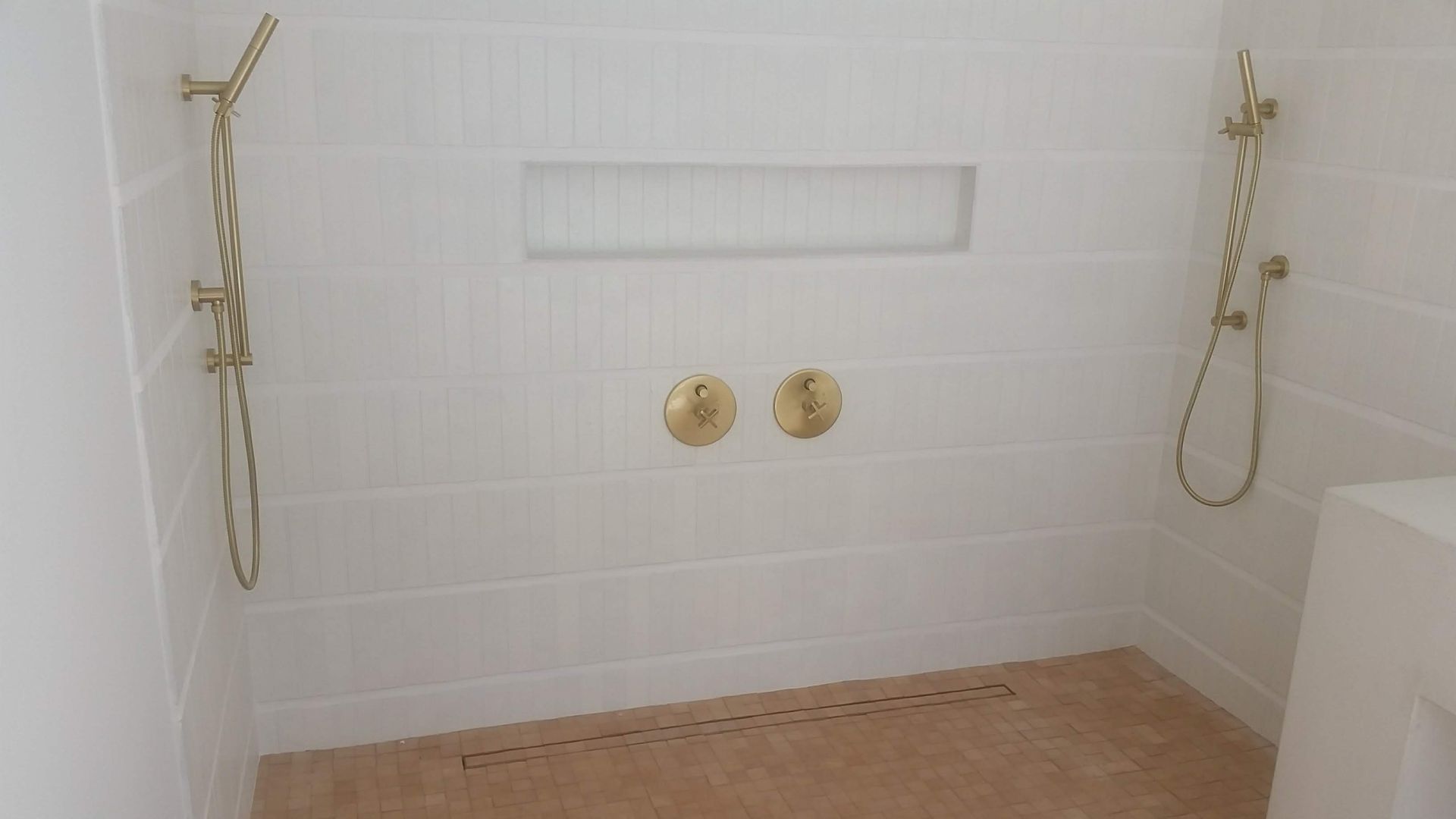 White tiled shower with gold fixtures, a built-in shelf, and textured tan floor.