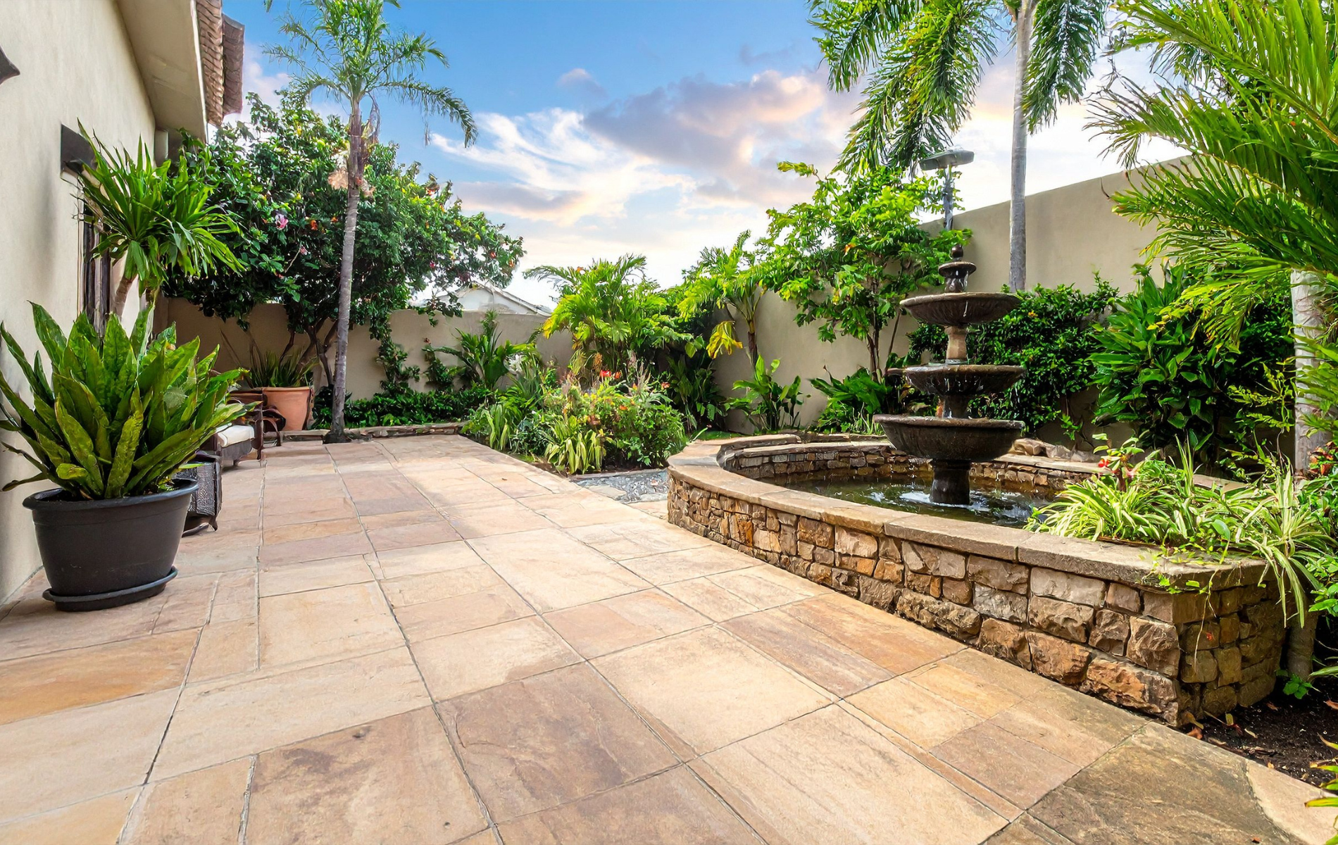Stone patio with fountain, surrounded by lush greenery and palm trees. Overcast sky.