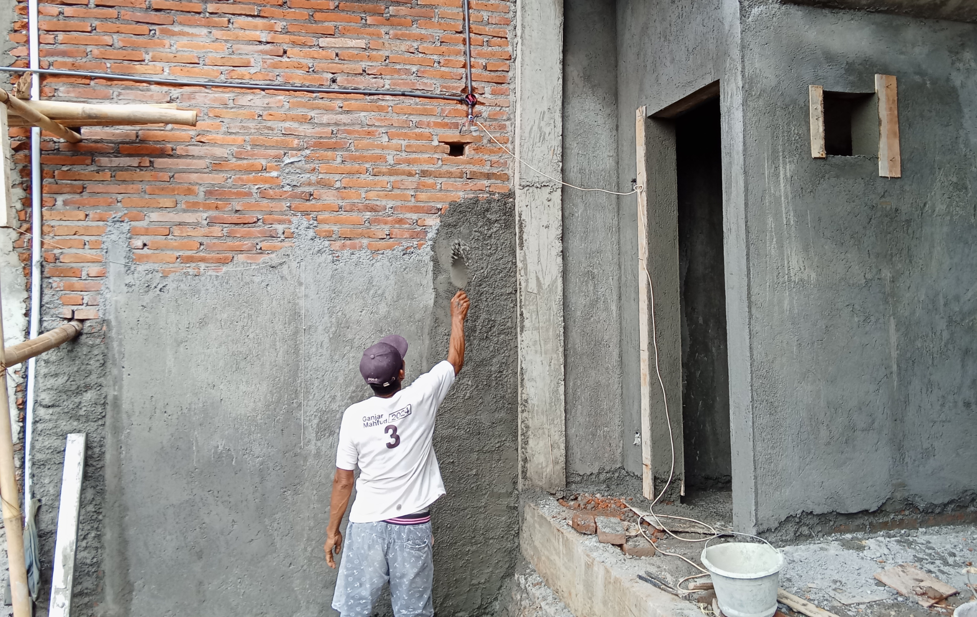 Construction worker plastering a gray concrete wall next to a brick wall with a wooden door frame.
