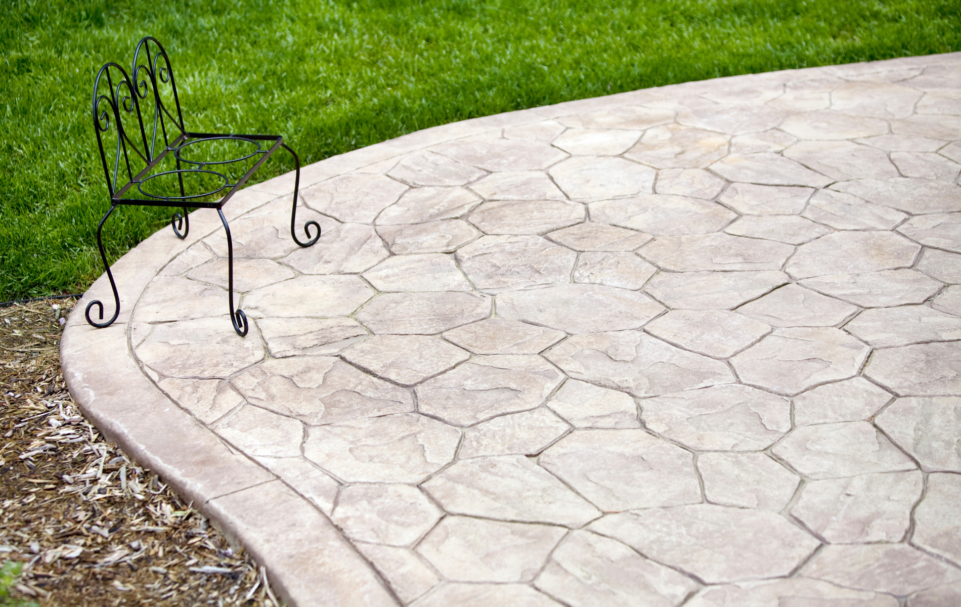 Stone-patterned concrete patio with a decorative black metal chair on the edge of green grass.