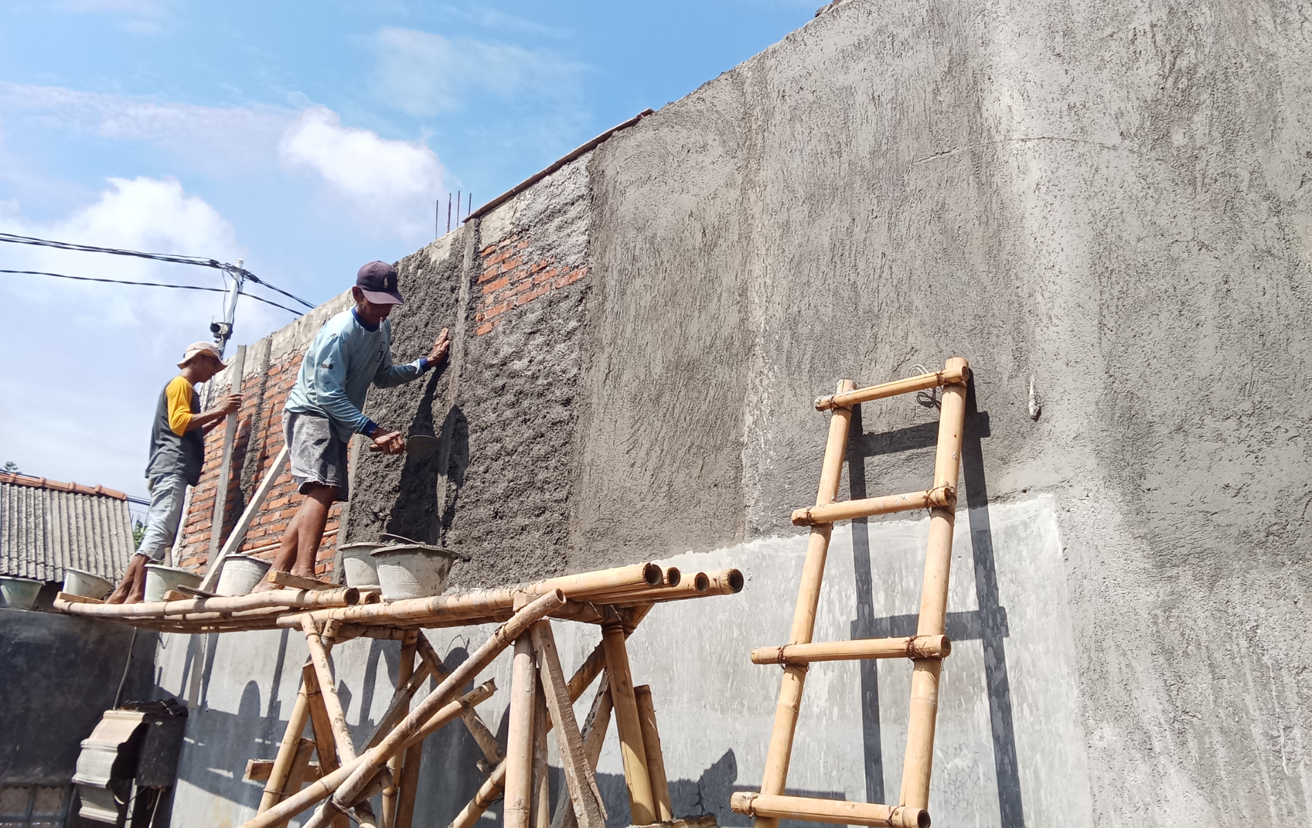 Two workers plastering a brick wall; one on a bamboo platform, another using a ladder, under a blue sky.