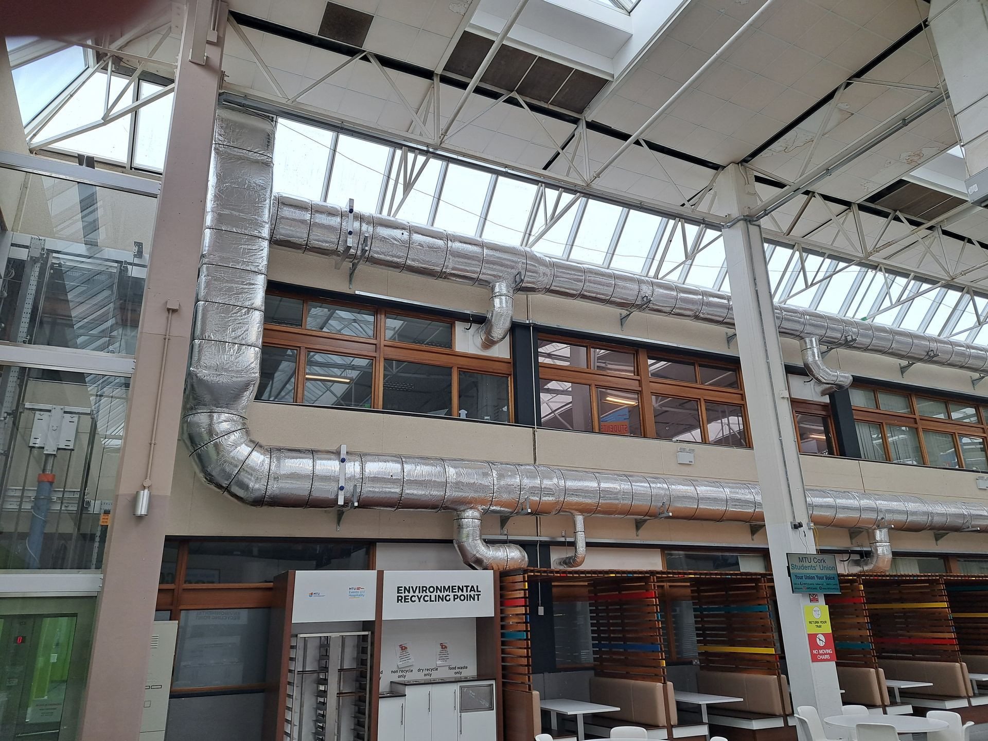 Interior view of a building with windows, ductwork, and open seating. Sunlight streams through the skylight.