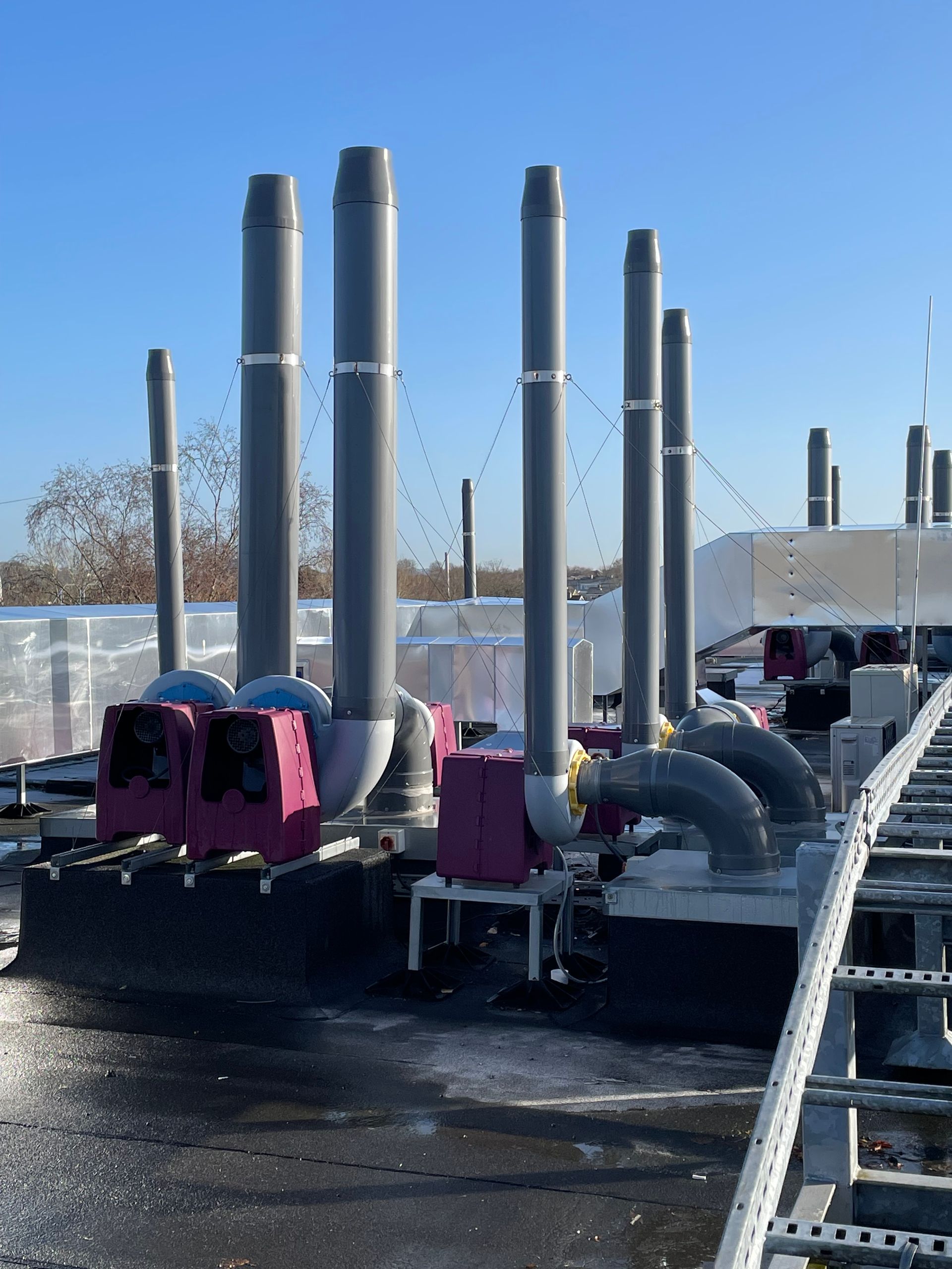 Grey industrial pipes and exhaust stacks on a roof against a clear blue sky.