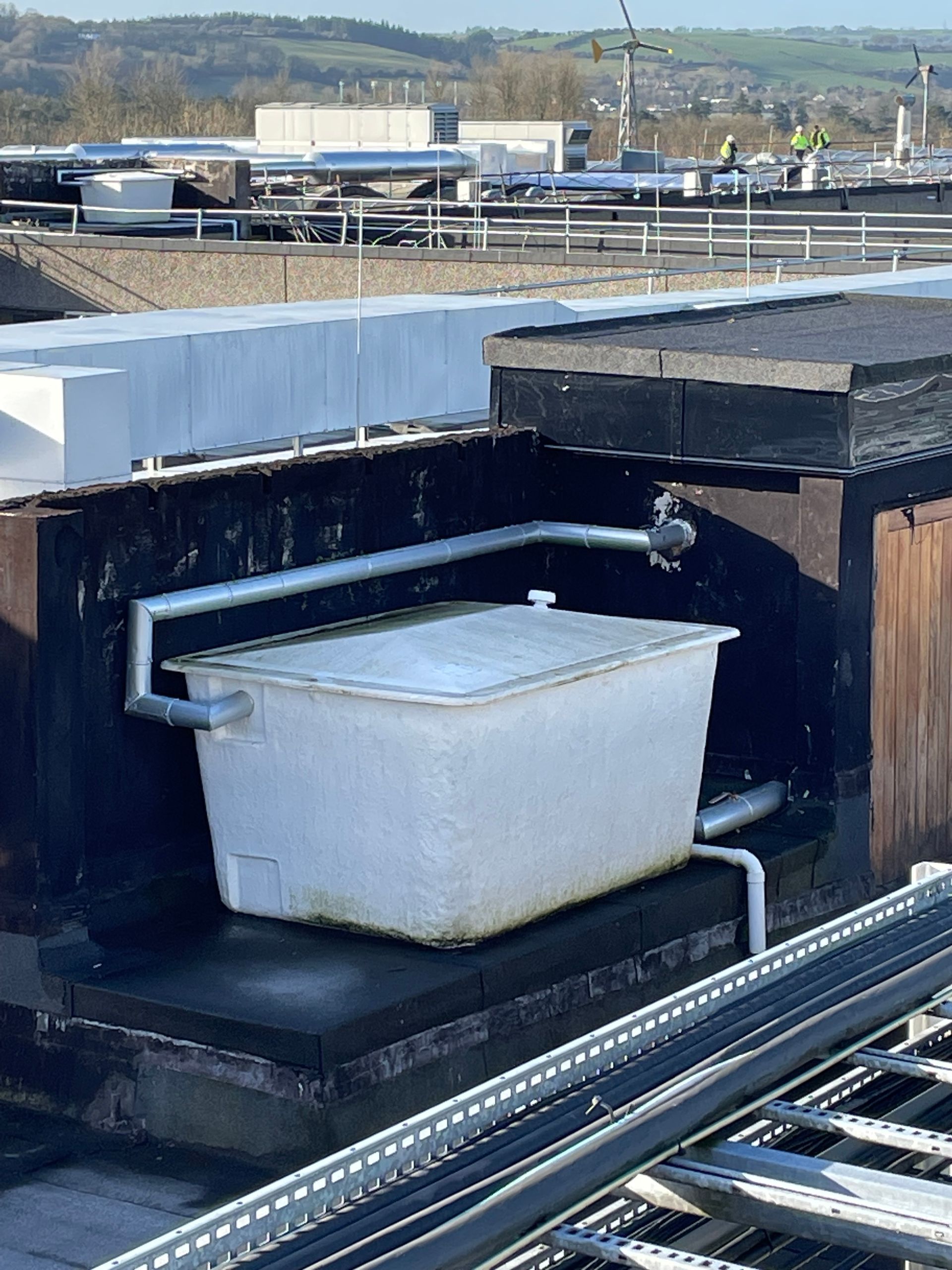 White water tank on a flat rooftop with pipes connected. In background, rooftop equipment and buildings.