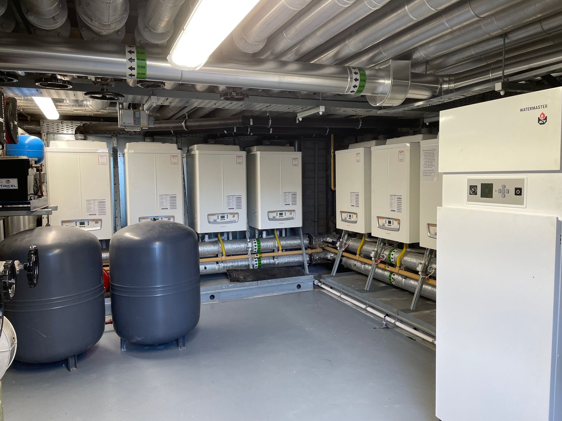 Heating system room with several white boilers, pipes, and tanks.