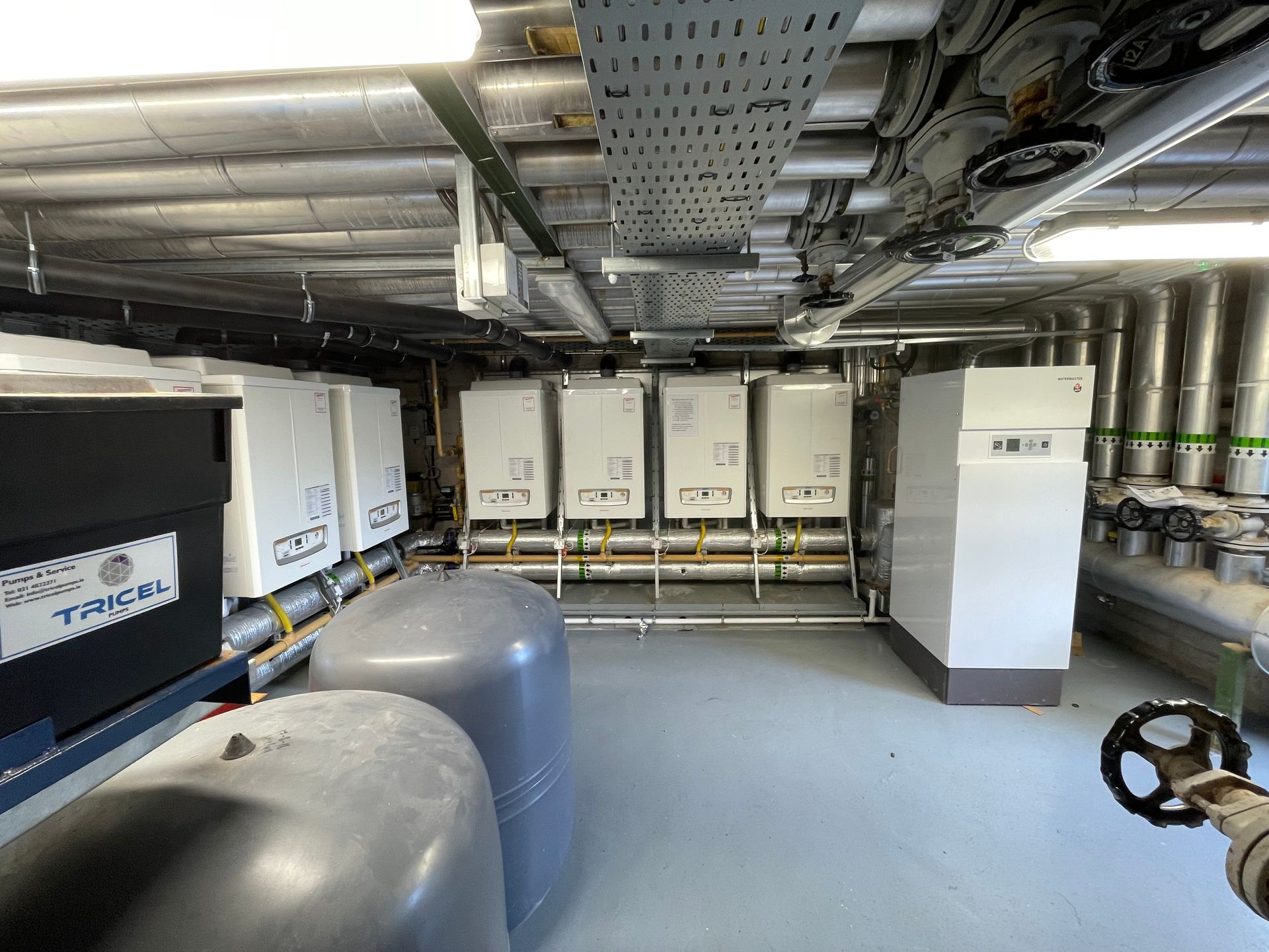 Industrial room with pipes, white machinery, and tanks on the floor.