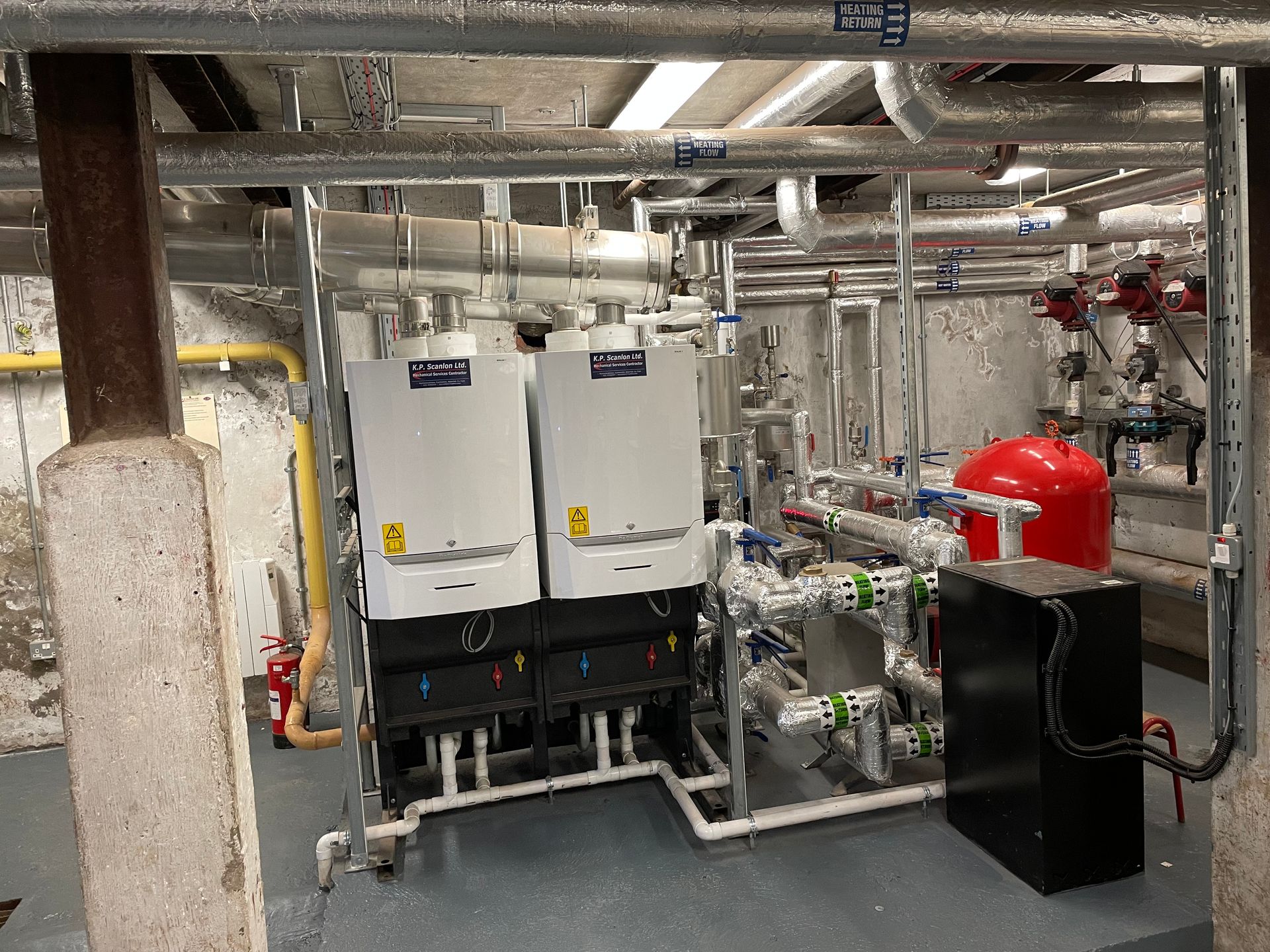 Heating system with two white boilers, red expansion tank, and pipes in an industrial setting.