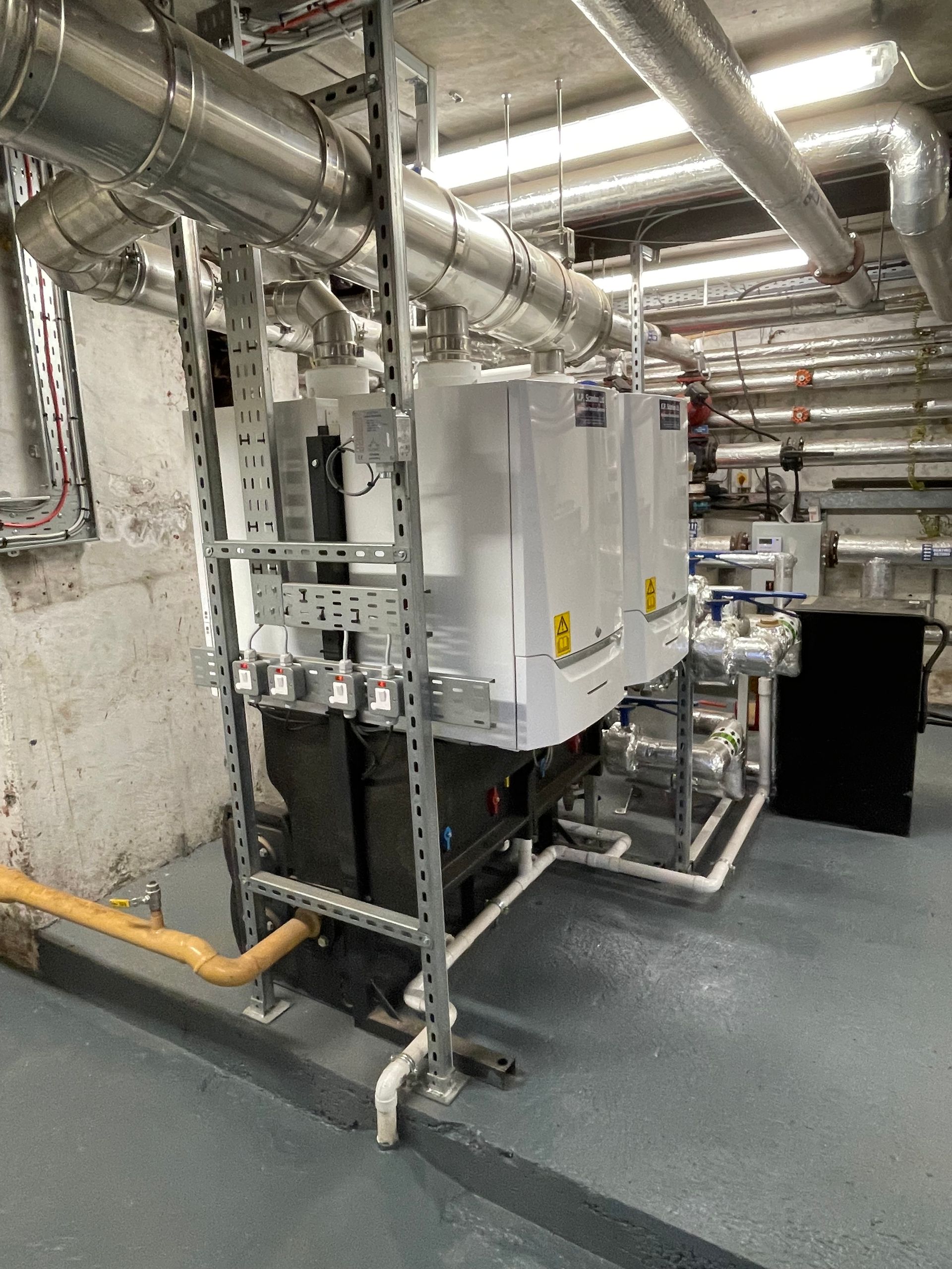 Heating system with silver pipes and white units in an industrial setting. Grey floor and concrete wall.
