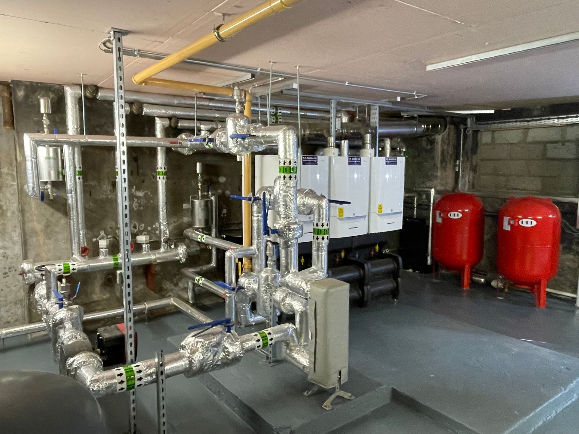 Heating system in a basement with insulated pipes, boilers, red tanks, and concrete walls.