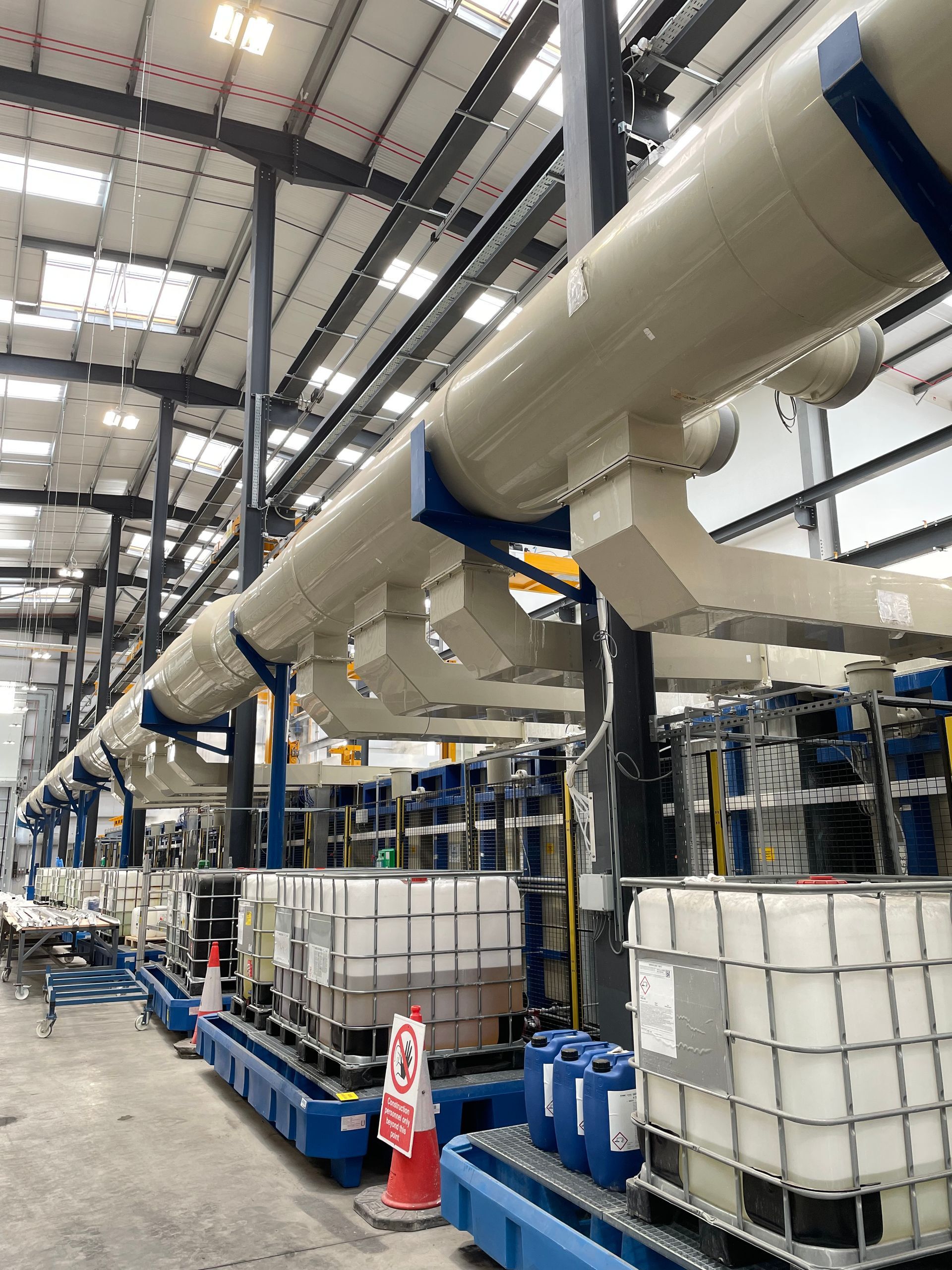 Industrial plant interior with large white pipes, chemical containers, and metal supports.