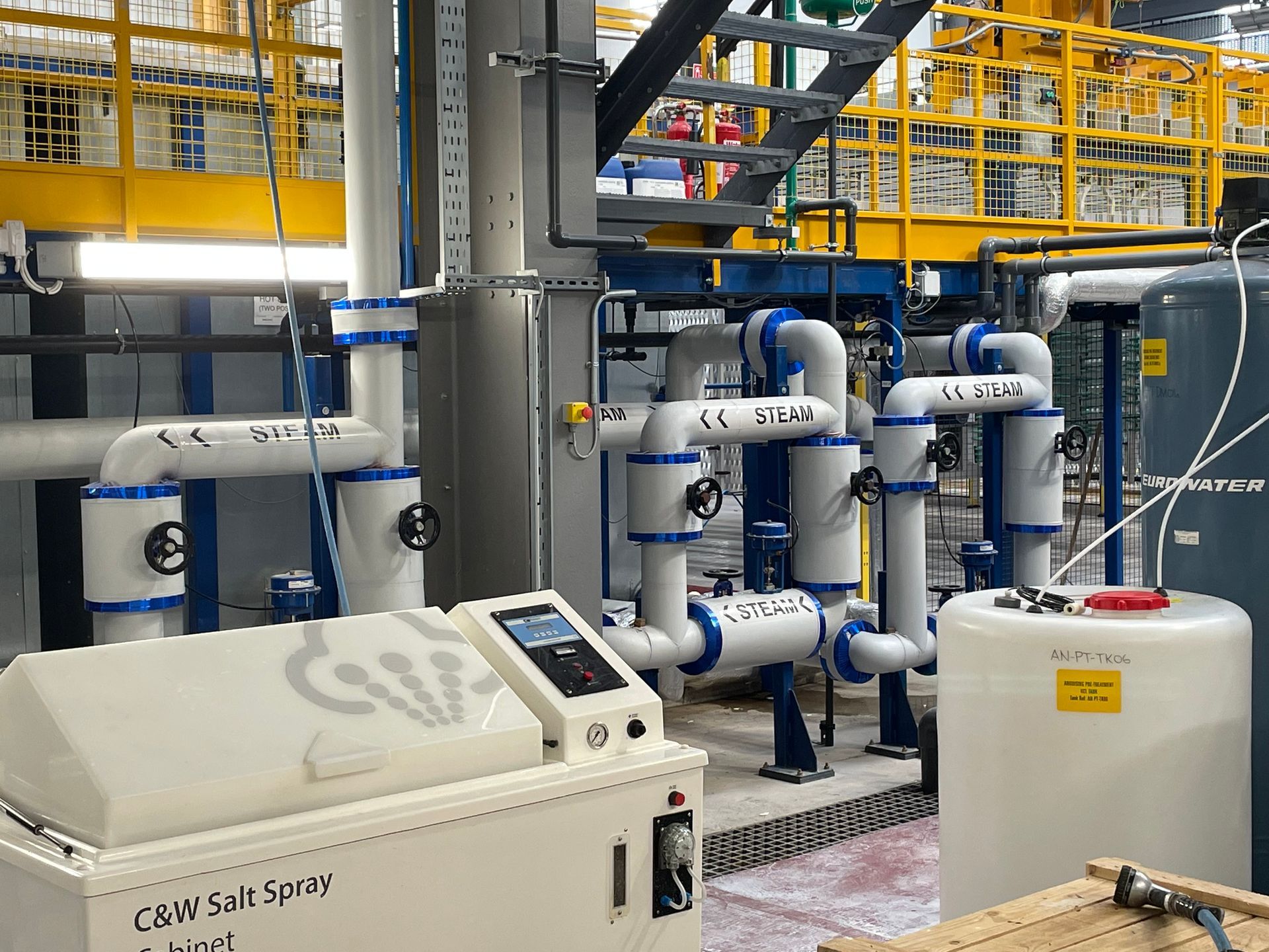 Industrial setting with pipes, tanks, and a control panel for a CREW Salt Spray chamber.