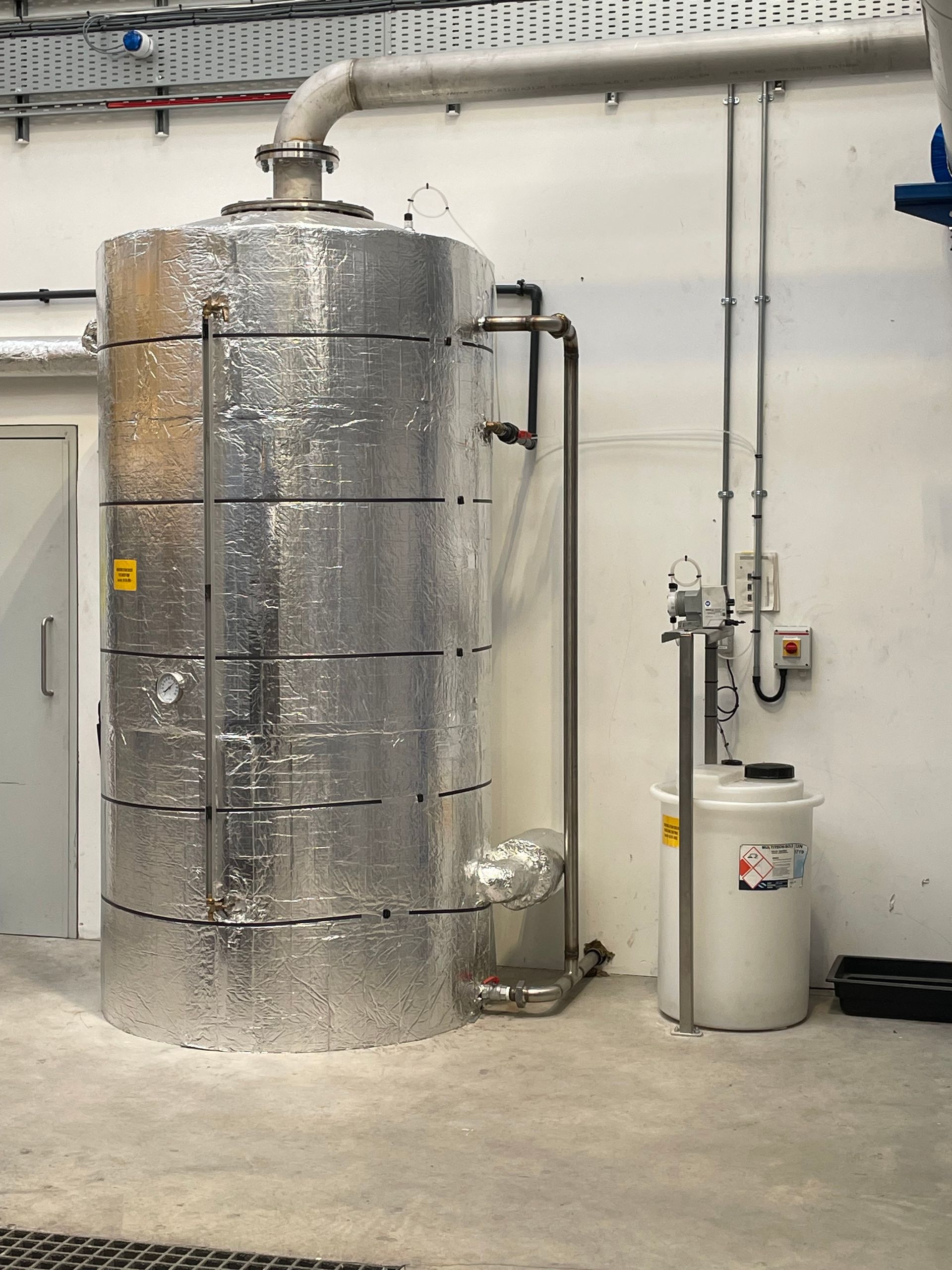 Silver insulated cylindrical tank in an industrial setting, with attached pipes and a white container nearby.