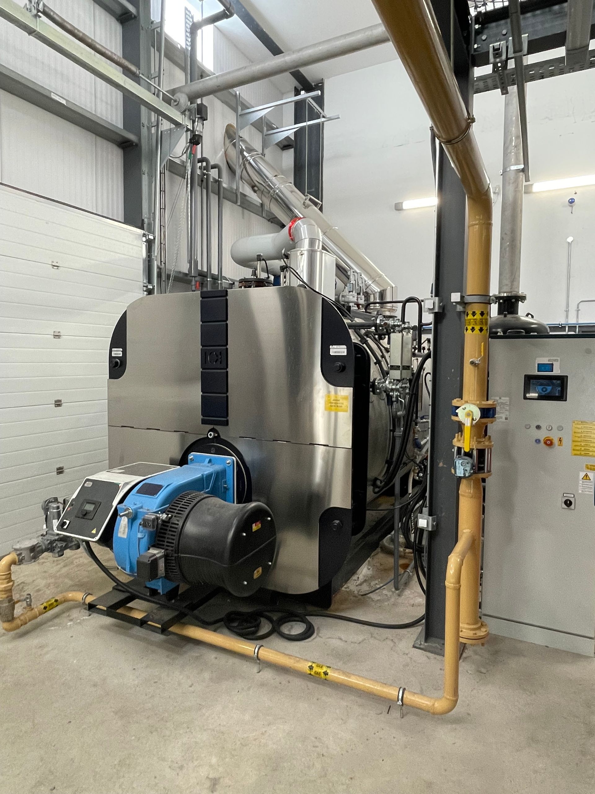 A large industrial boiler in a light-colored room. It has a blue burner and stainless steel casing with yellow pipes.