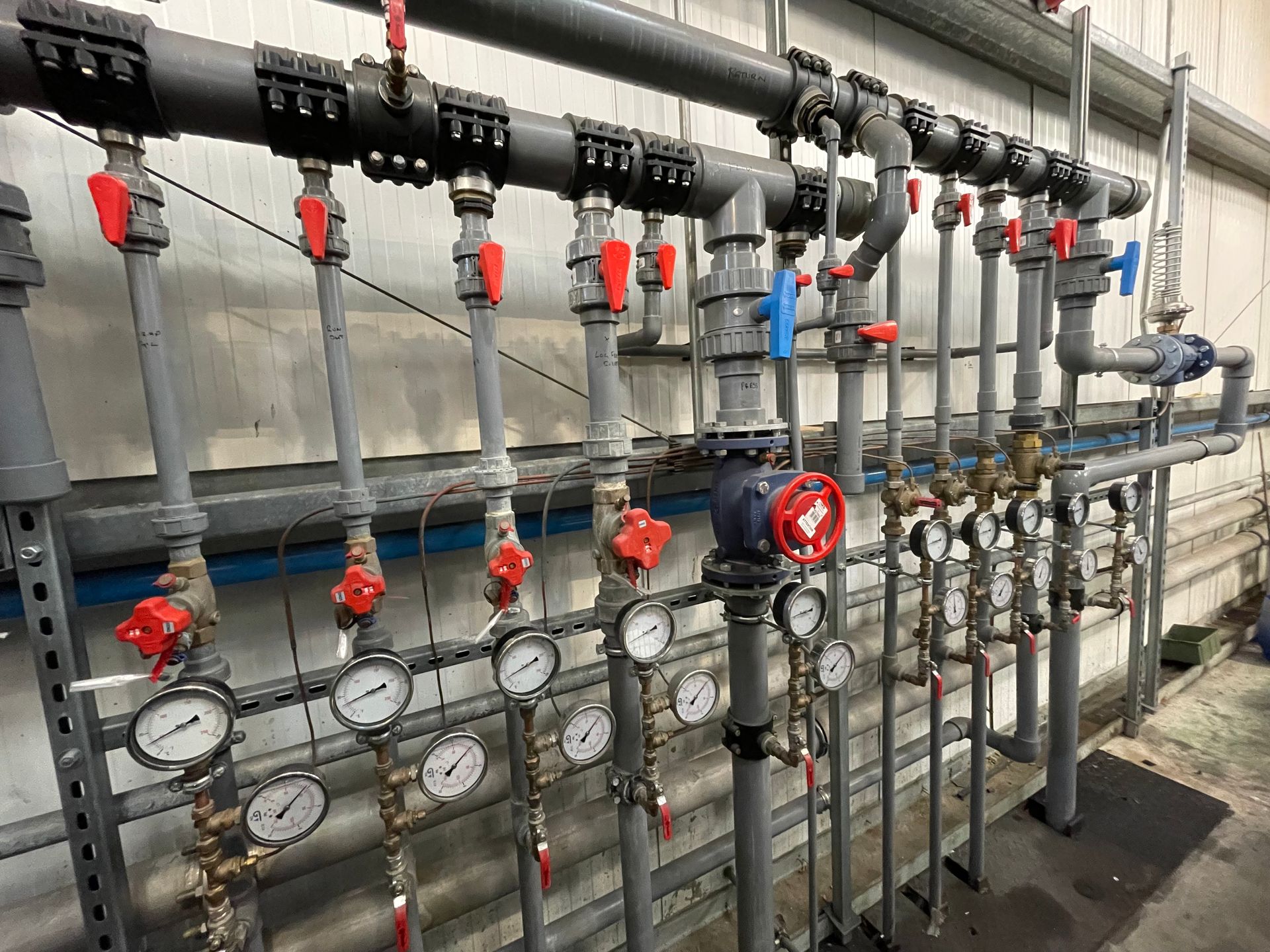 Piping system with valves, gauges, and red handles. Gray pipes and a metal rack against a wall.