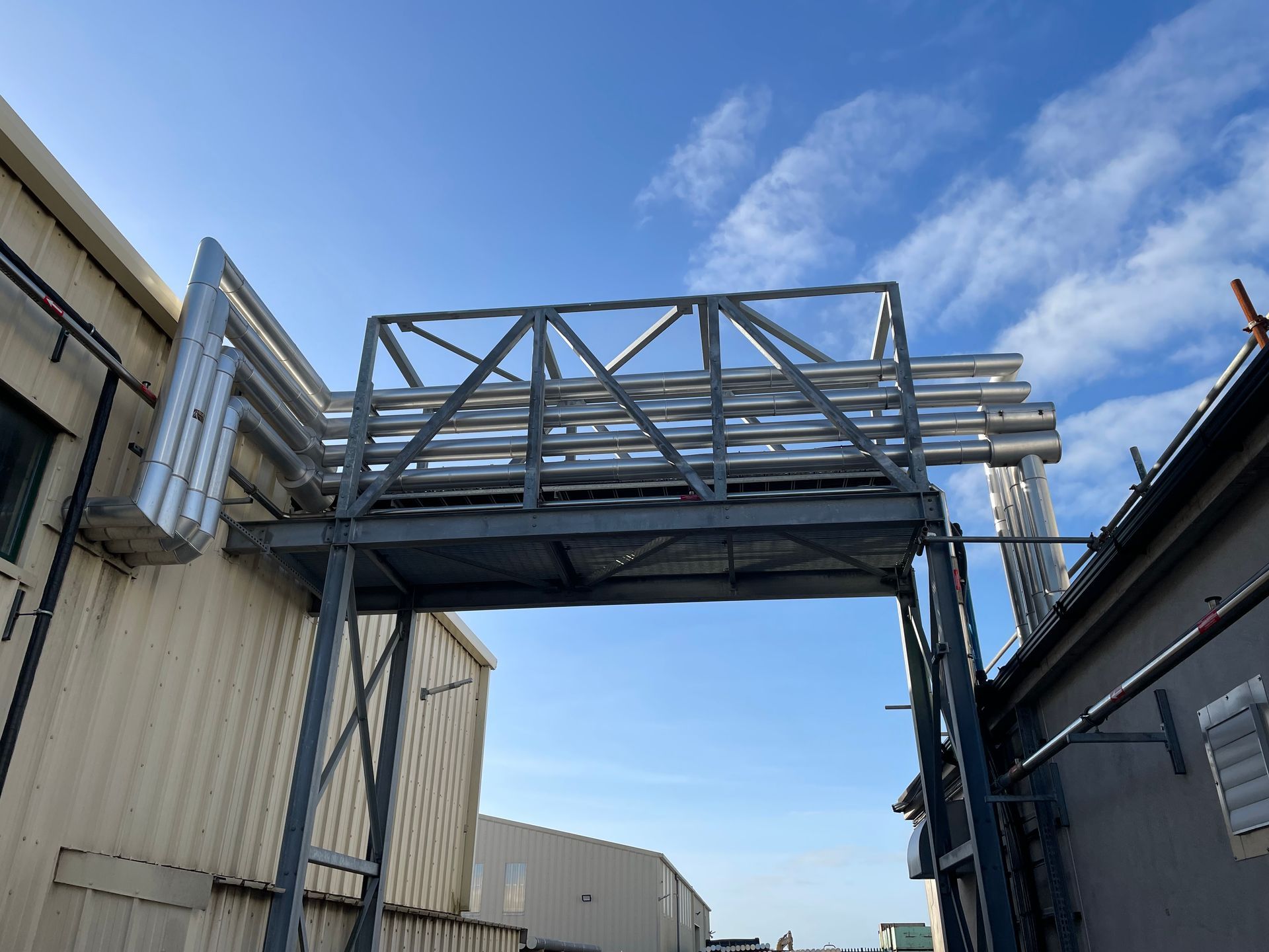 Metal bridge structure over pipes between two buildings against a cloudy blue sky.