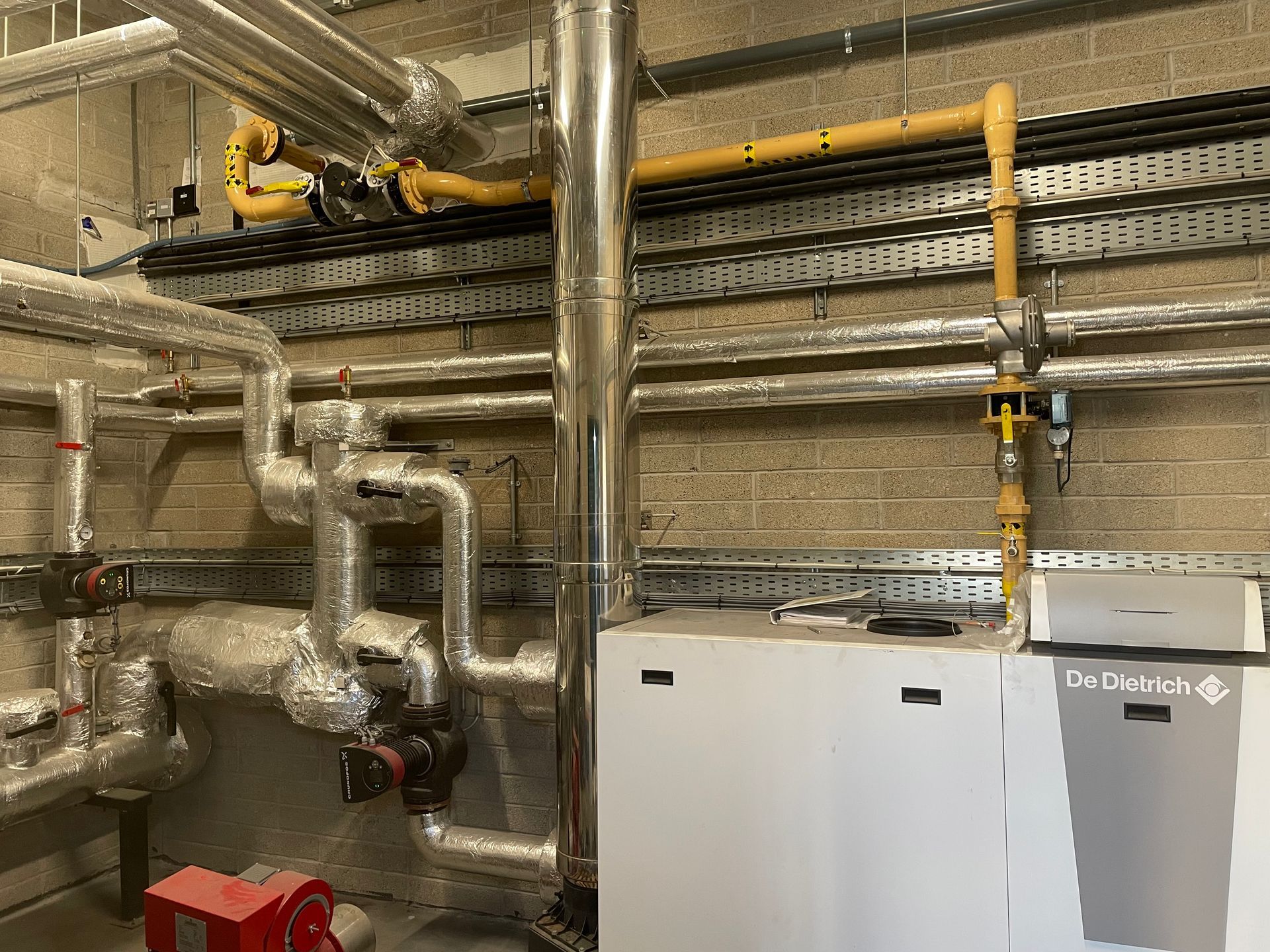 Pipes and mechanical equipment, possibly for heating or cooling, are mounted on a brick wall.