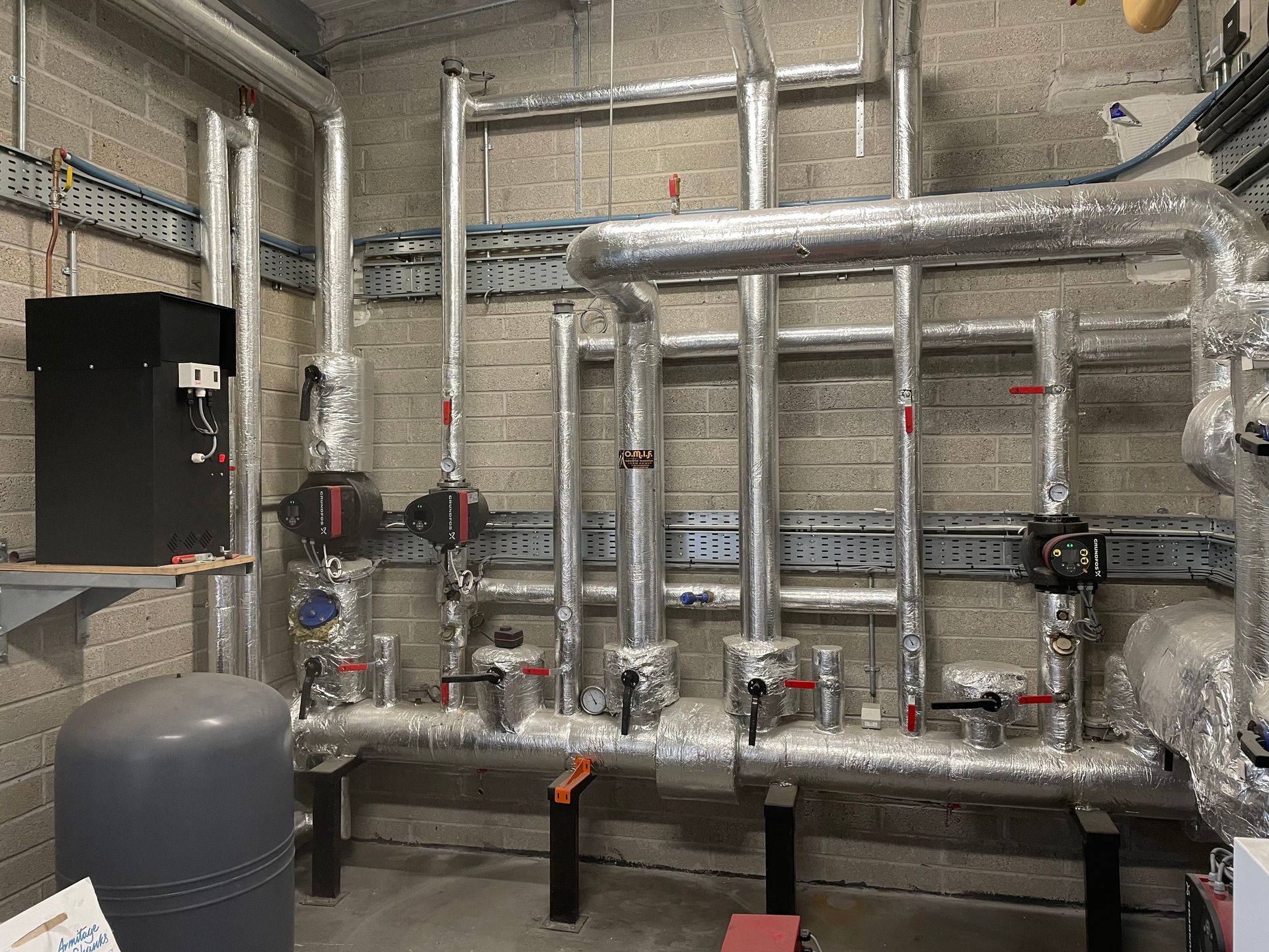 Pipes and equipment in an industrial setting, insulated with silver foil against a gray brick wall.