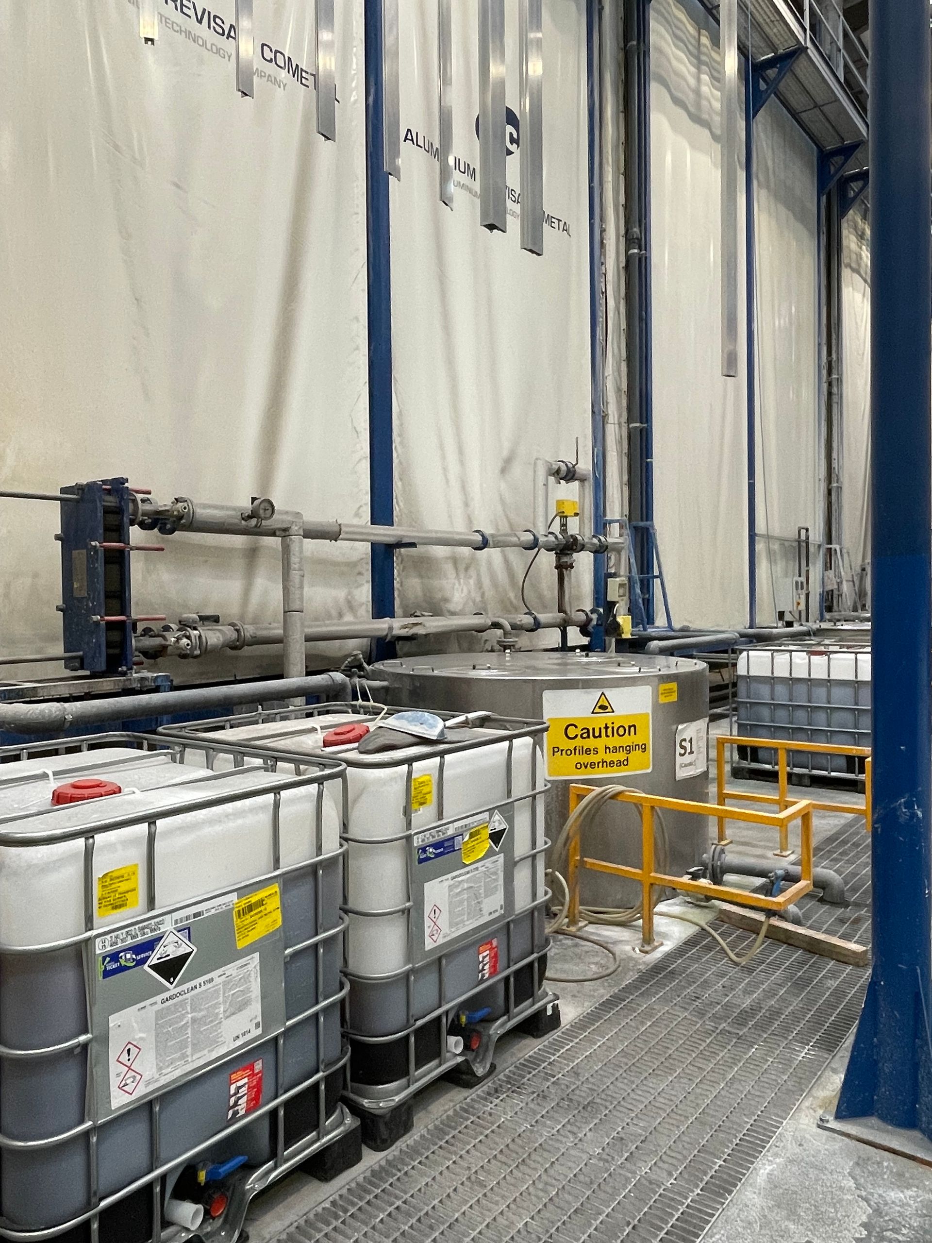 Industrial setting with tanks, pipes, and metal pieces hanging, likely for processing or finishing.