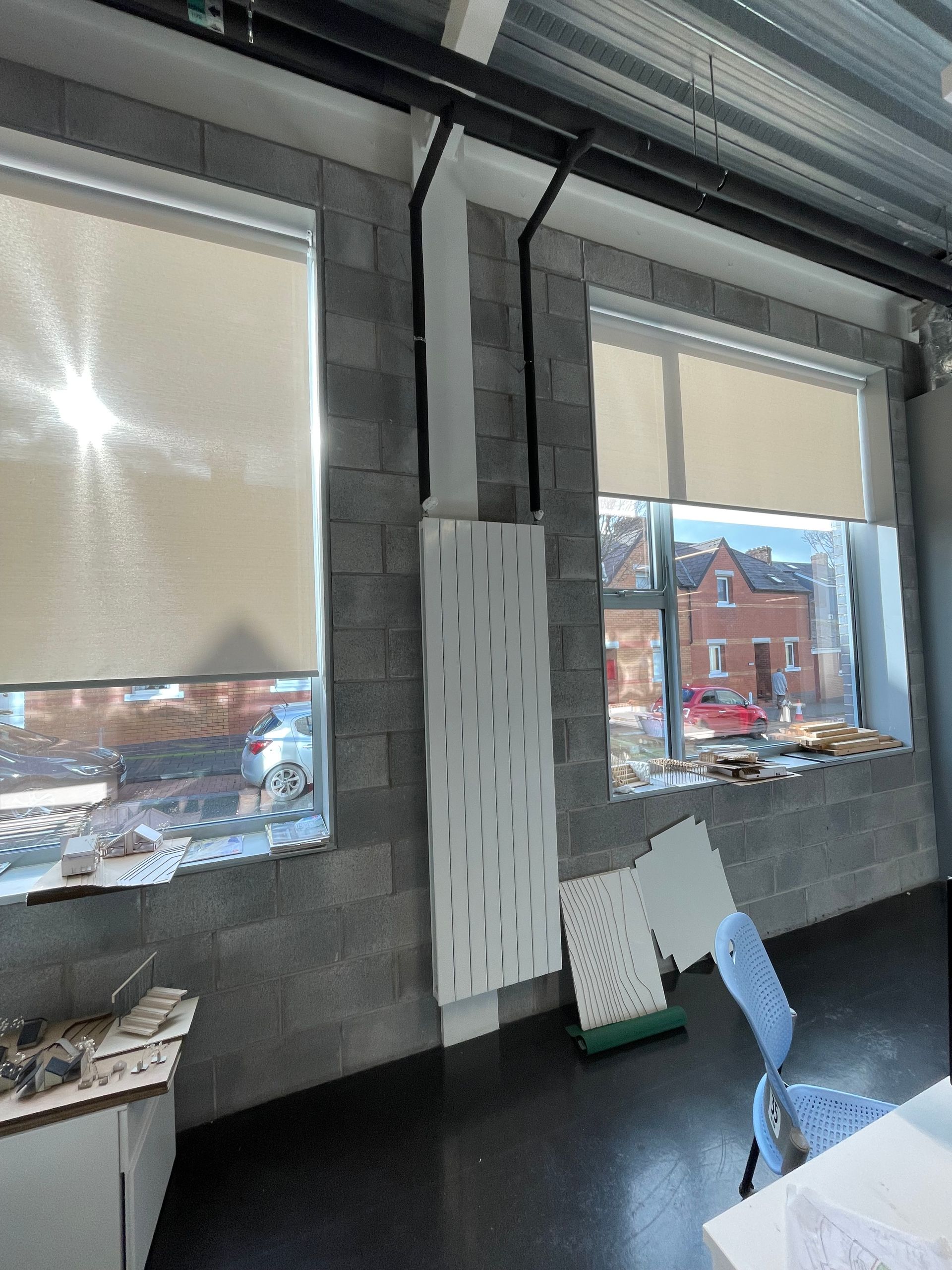 Two windows with beige blinds and cityscape views, gray brick wall with a white ventilation system, and black pipes.