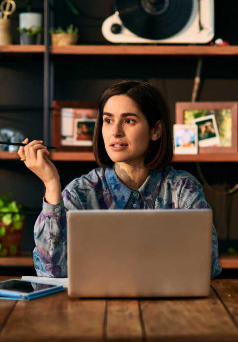 Woman at a desk with laptop, looking up and to the left, holding a pen. Indoor setting with wooden shelves and plants.