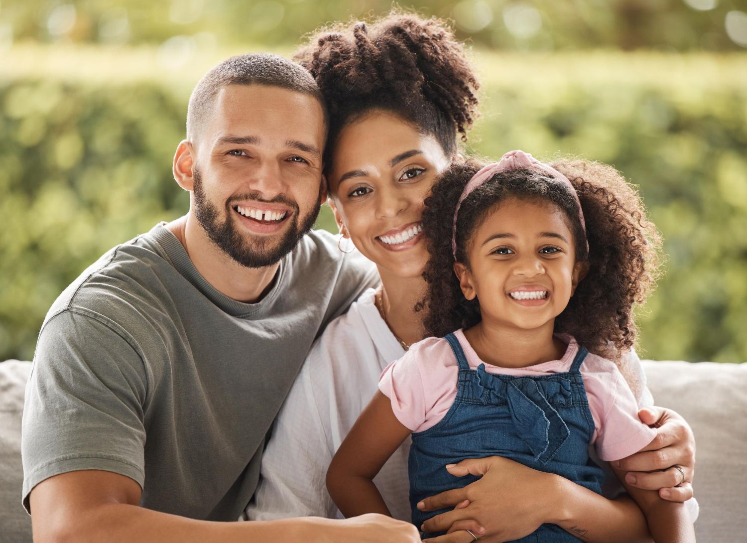 Happy family of three smiling: man, woman, and young girl, outdoors on a sunny day.