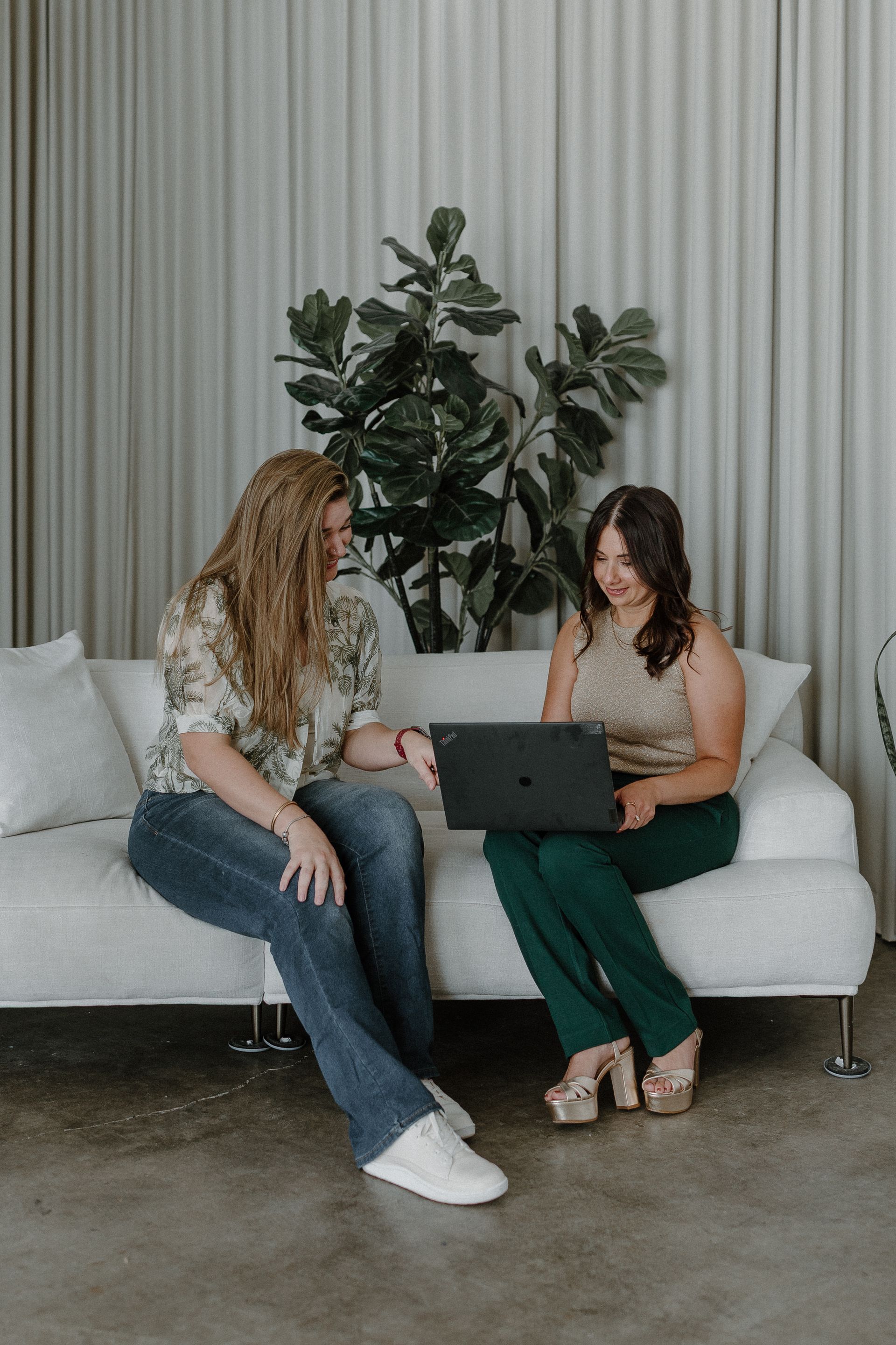 Woman in a white cardigan sits, smiling, in a bright setting; wearing a black top and gold necklace.