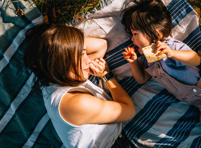 Woman and child lying on a striped blanket outdoors, child eating, woman looking at child.